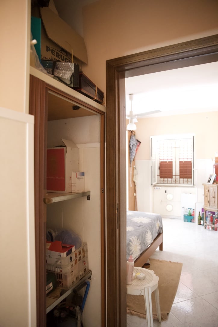 Scaffalatura in cucina con scatole e oggetti vari, vista su camera da letto con letto e finestra luminosa, interior design abitazione, appartamento accogliente e funzionale, stile semplice e pratico.