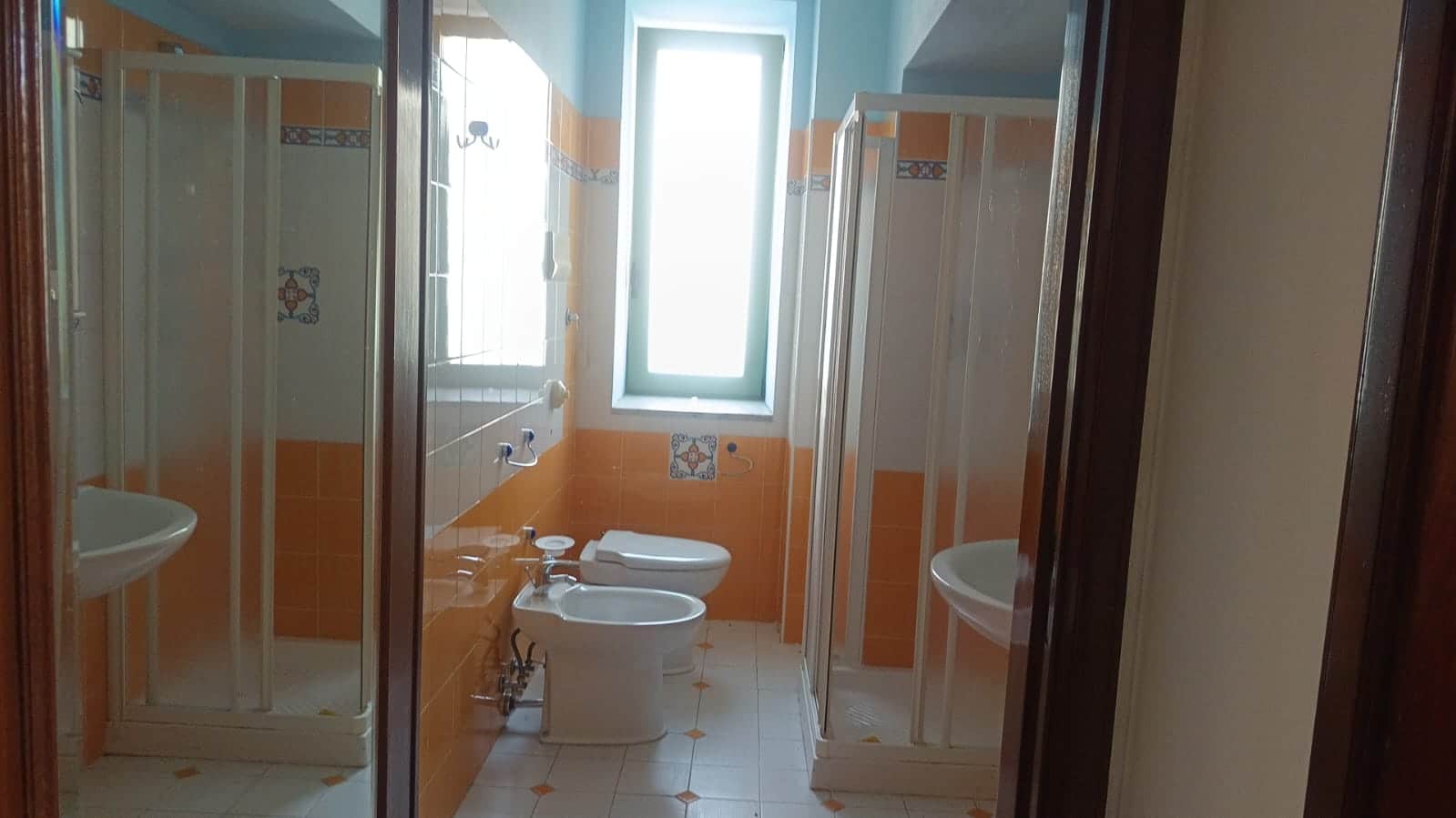 Servizio igienico con doccia, WC e bidet in stile classico, illuminato da finestra grande, in bagno con piastrelle arancioni e bianche. Ideale per case in vendita o affitto, con dettagli di design tradizionale.