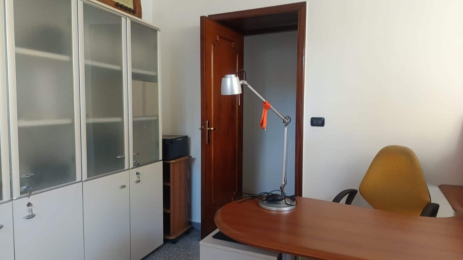 Immagine di un ufficio con pareti chiare, armadiature con ante in vetro, scrivania in legno e sedia gialla, lampada da scrivania regolabile, atmosfera professionale e funzionale.