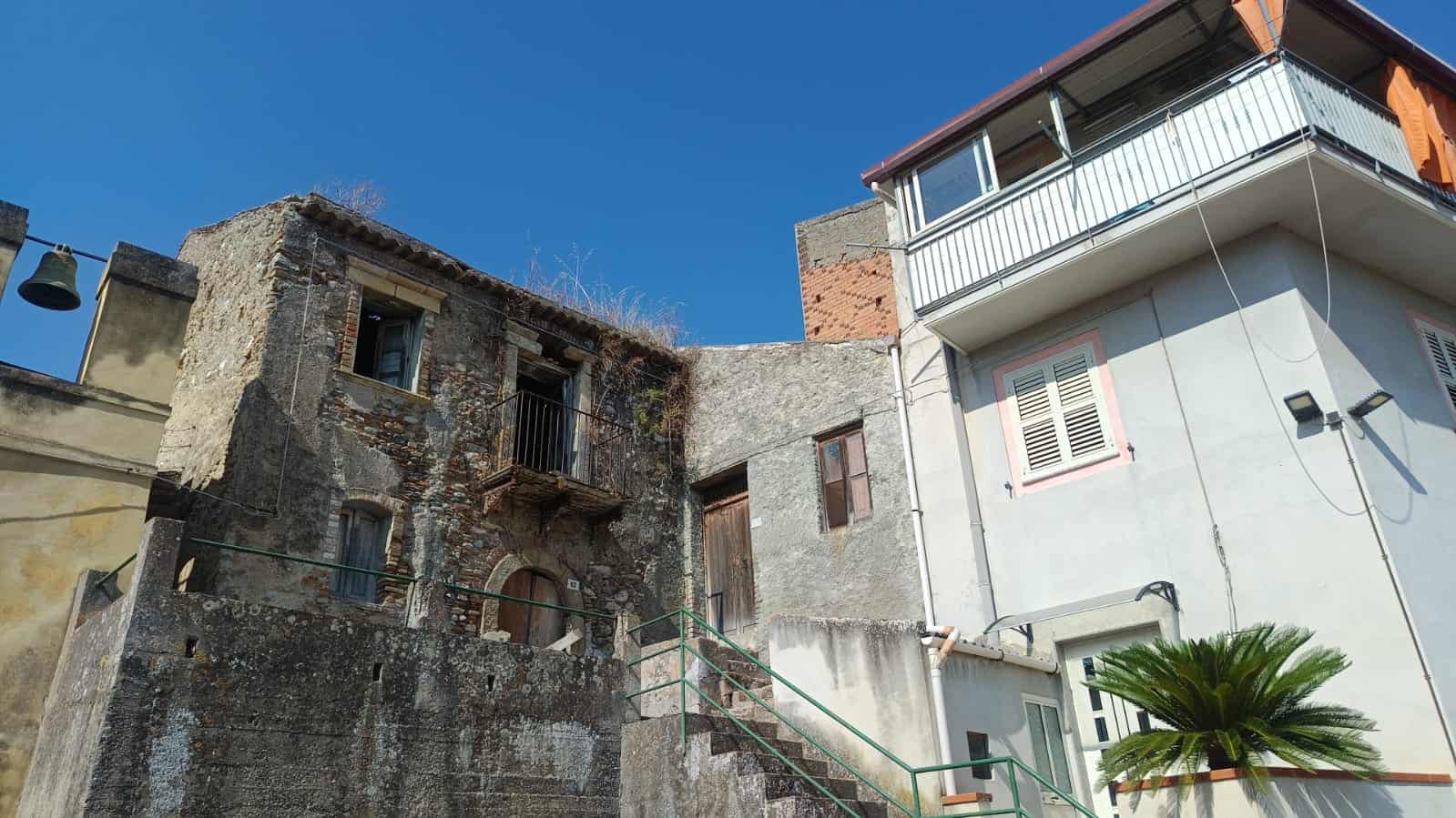 f1cdc174-4c56-4019-8ddb-07a0edf9b445 Vecchie case in stile rustico e moderna, con scale esterne e balcone, ambientazione urbana, cielo azzurro di giorno.