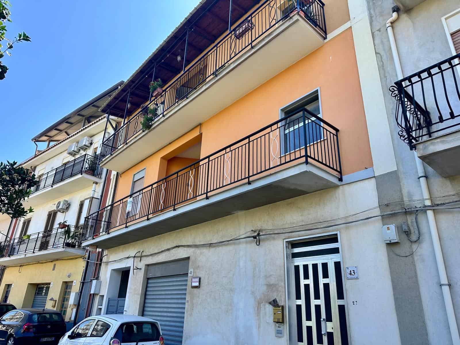Balcone di un appartamento in edificio residenziale con facciata colorata, ringhiere in ferro battuto, strada con auto parcheggiate, situato in zona urbana con clima soleggiato.
