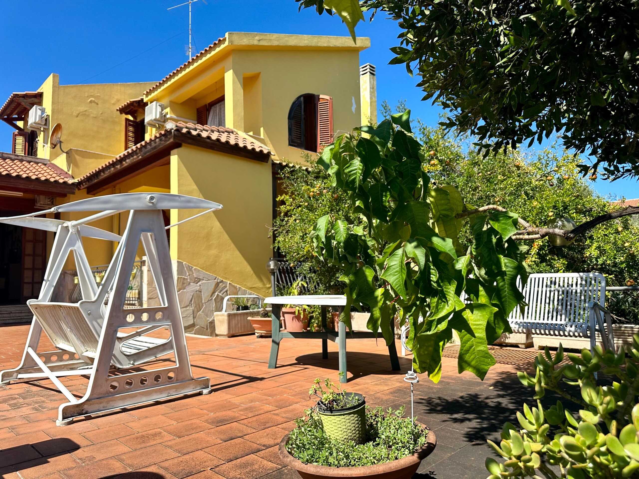 Vista di un terrazzo soleggiato con mobili da giardino, piante verdi e un edificio residenziale giallo con tetto di tegole, ideale per l'acquisto o l'affitto di case tramite NextCasa.