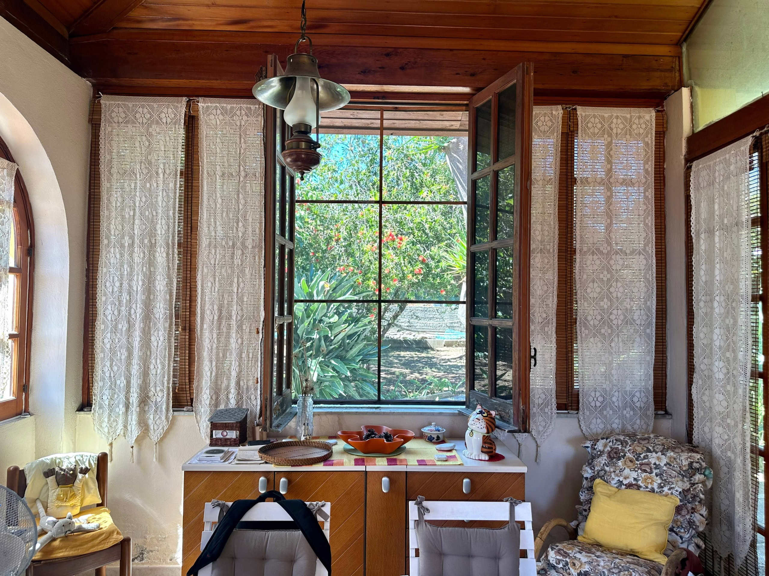 Finestra aperta su un giardino rigoglioso con tende in pizzo, interni accoglienti e decorazioni vintage in una casa rustica. Perfetto per chi cerca un'atmosfera calda e naturale.