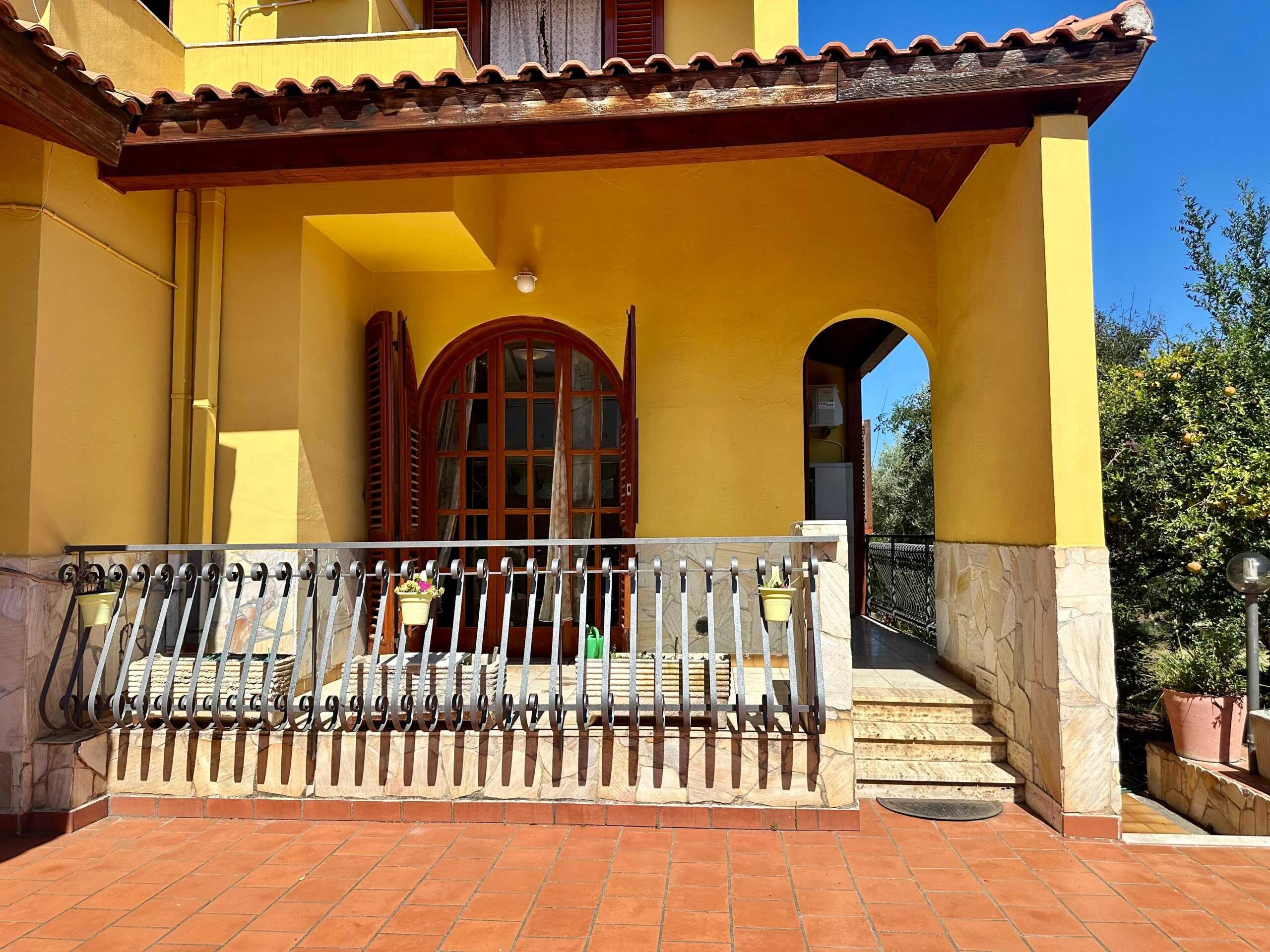 Accogliente terrazza di casa gialla con ringhiera in ferro e piante in vaso, ideale per relax all'aperto e momenti conviviali.