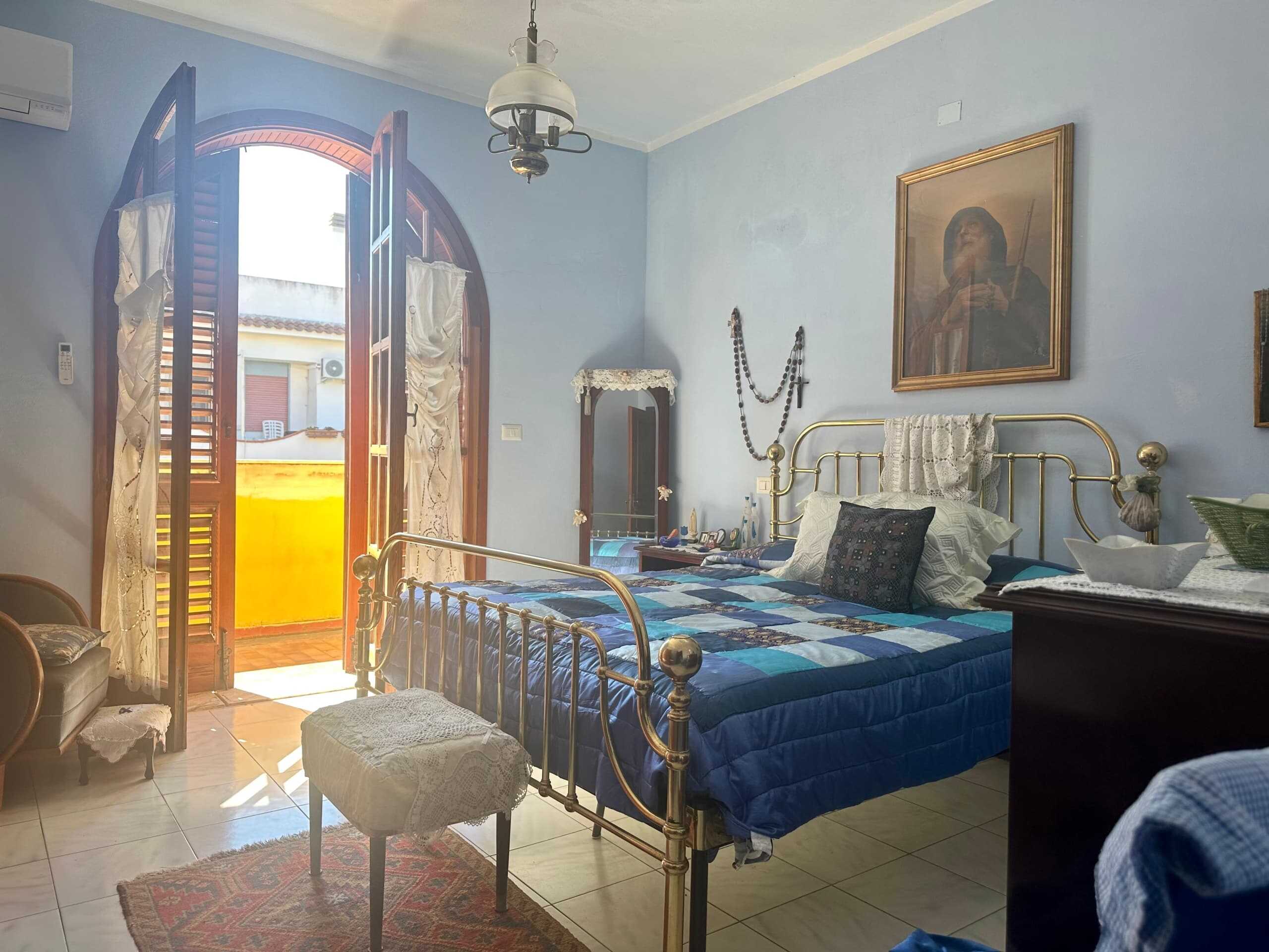 Balcone al sole in camera da letto con affaccio su edificio residenziale, arredamento classico, mobili in legno e dettagli decorativi vintage, luce naturale diffusa, atmosfera accogliente e tranquilla.