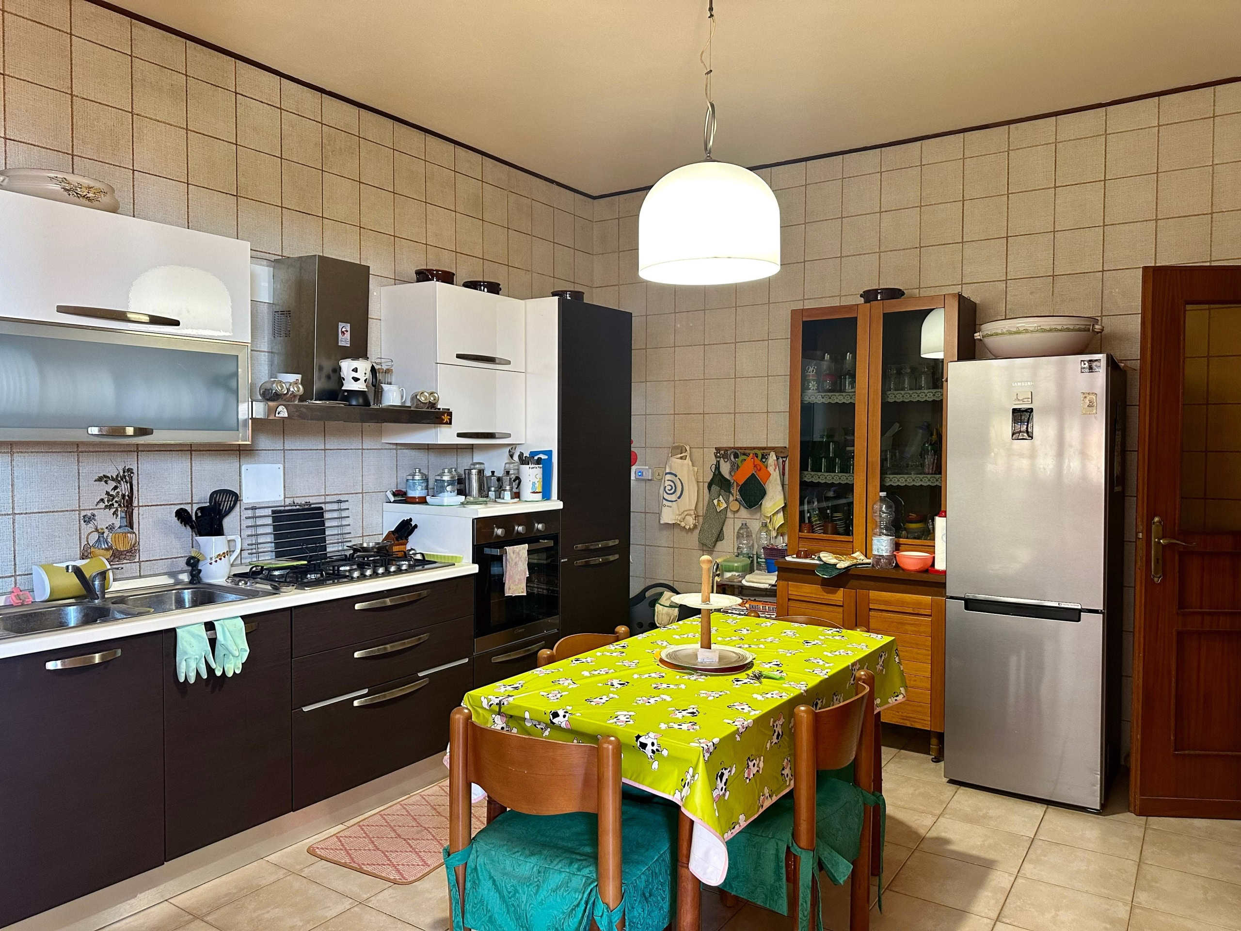 Camino in cucina con mobili moderni, tavolo con tovaglia a tema mucche, frigorifero in acciaio, stoviglie e utensili, ambiente funzionale e accogliente.