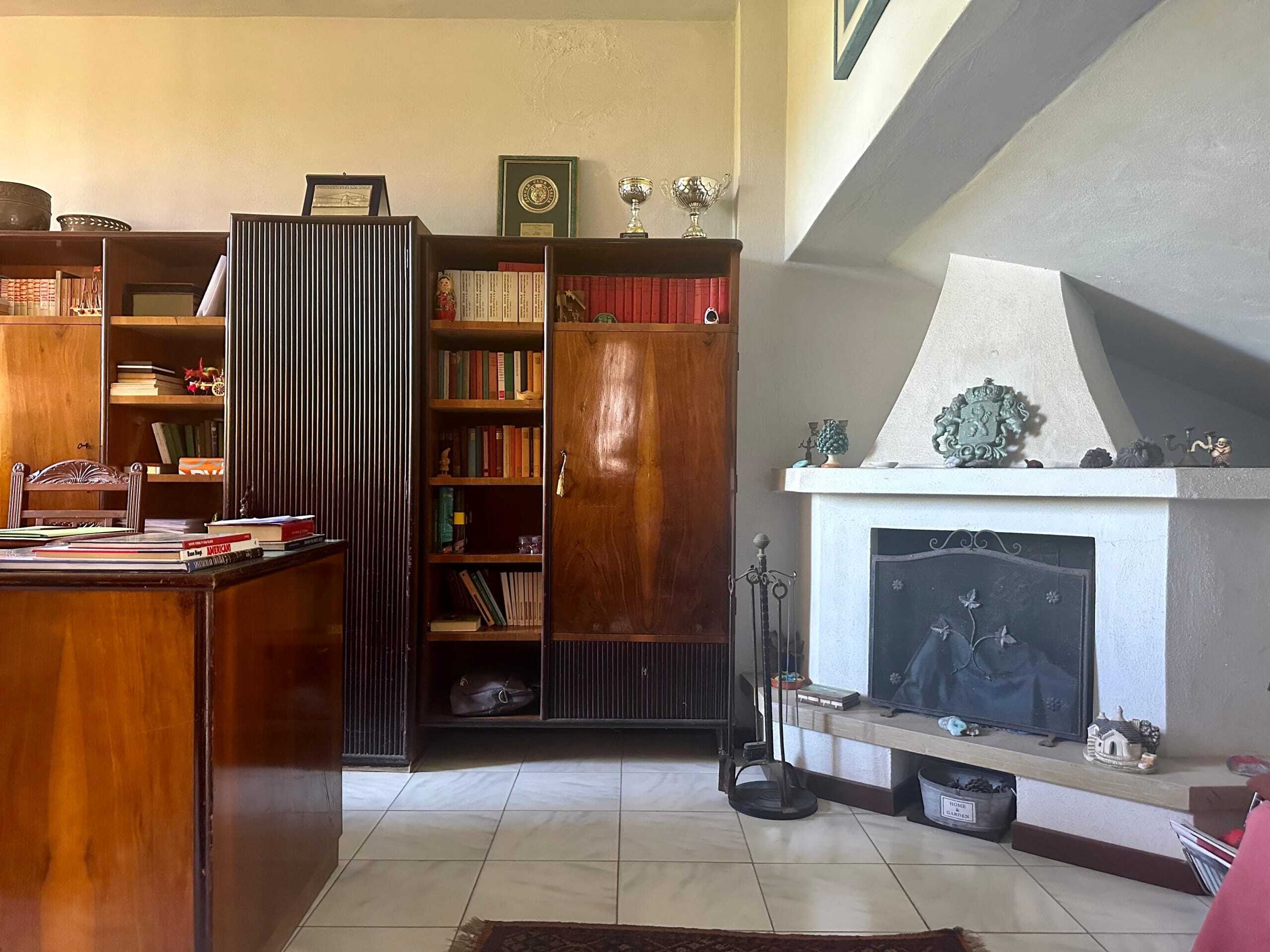 Esterni di una casa con pareti bianche, camino decorato, scaffali in legno ricchi di libri e oggetti da collezione, atmosfera accogliente e tradizionale.