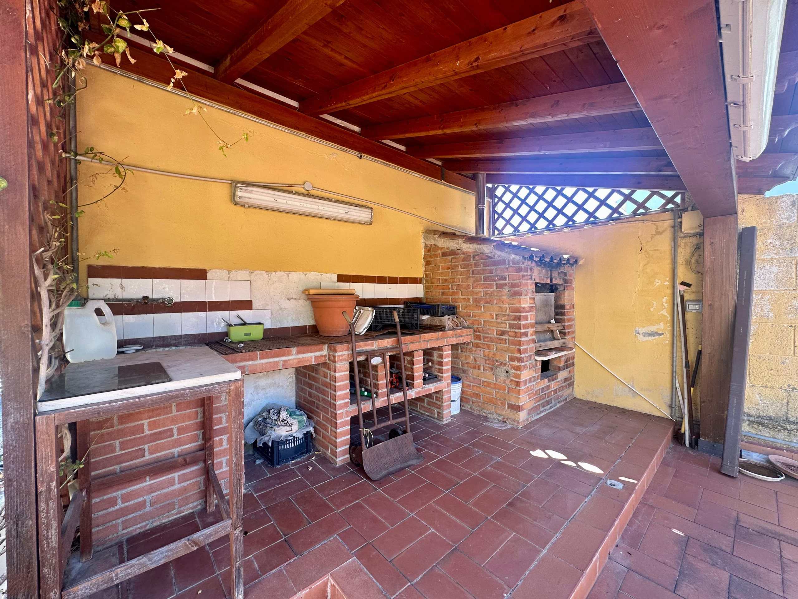 Spazio esterno con cucina all'aperto e barbecue in casa NextCasa, ideale per pranzi e cene estive, dotata di mattoni, piante e zona coperta in legno.