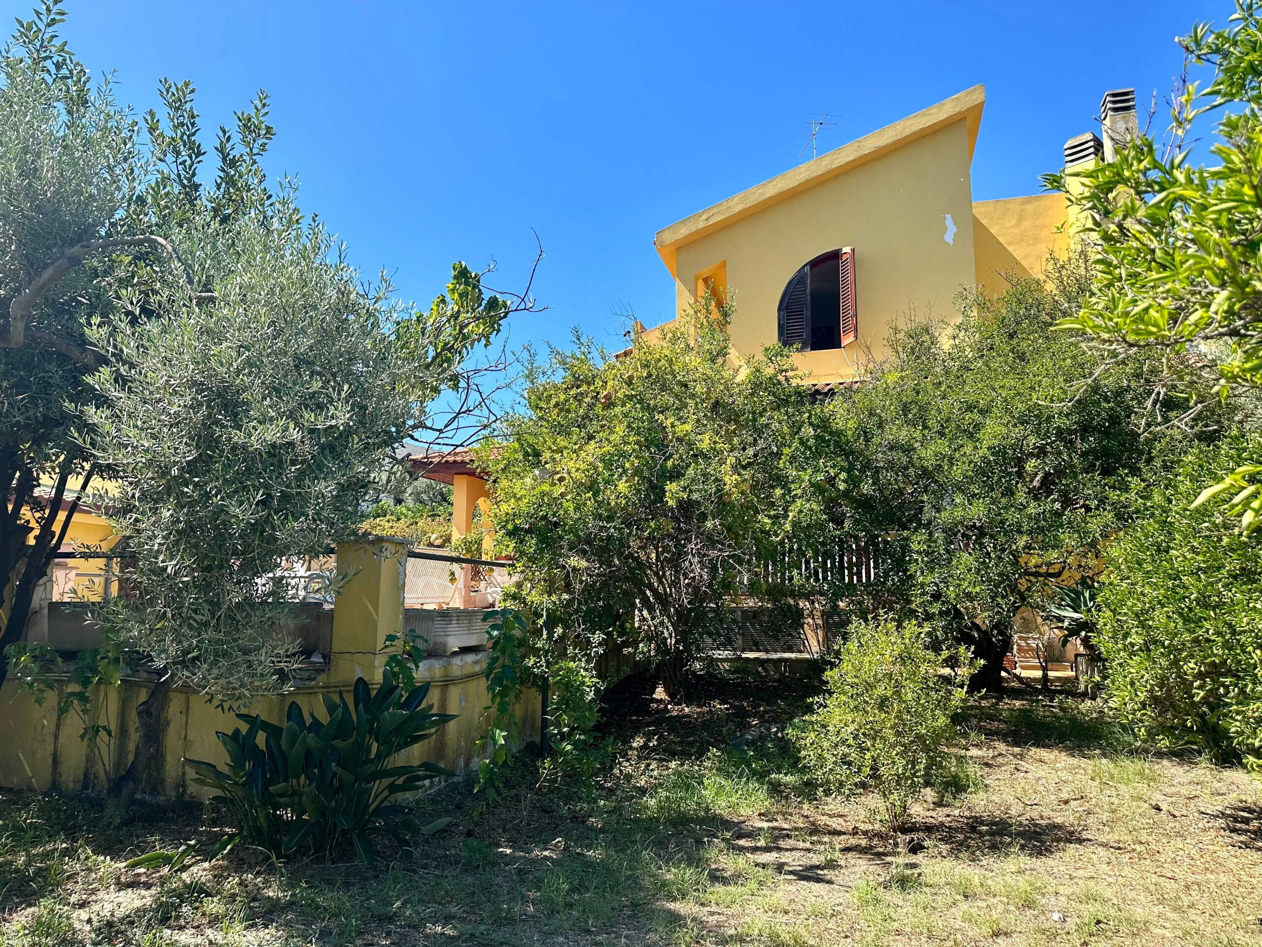 Immagine di una casa tradizionale in stile mediterraneo circondata da alberi verdi e piante ornamentali, con una facciata gialla e finestre con persiane scure, sotto un cielo azzurro brillante.