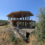 Impalcatura di un edificio in costruzione con struttura in cemento armato, senza pareti, su un terreno collinare con vegetazione naturale e vista panoramica.