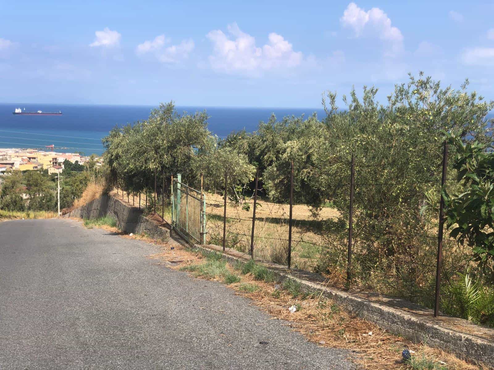 Vista panoramica sul mare e paesaggio collinare, strada e recinzione in una zona residenziale vicino al mare in Sicilia.