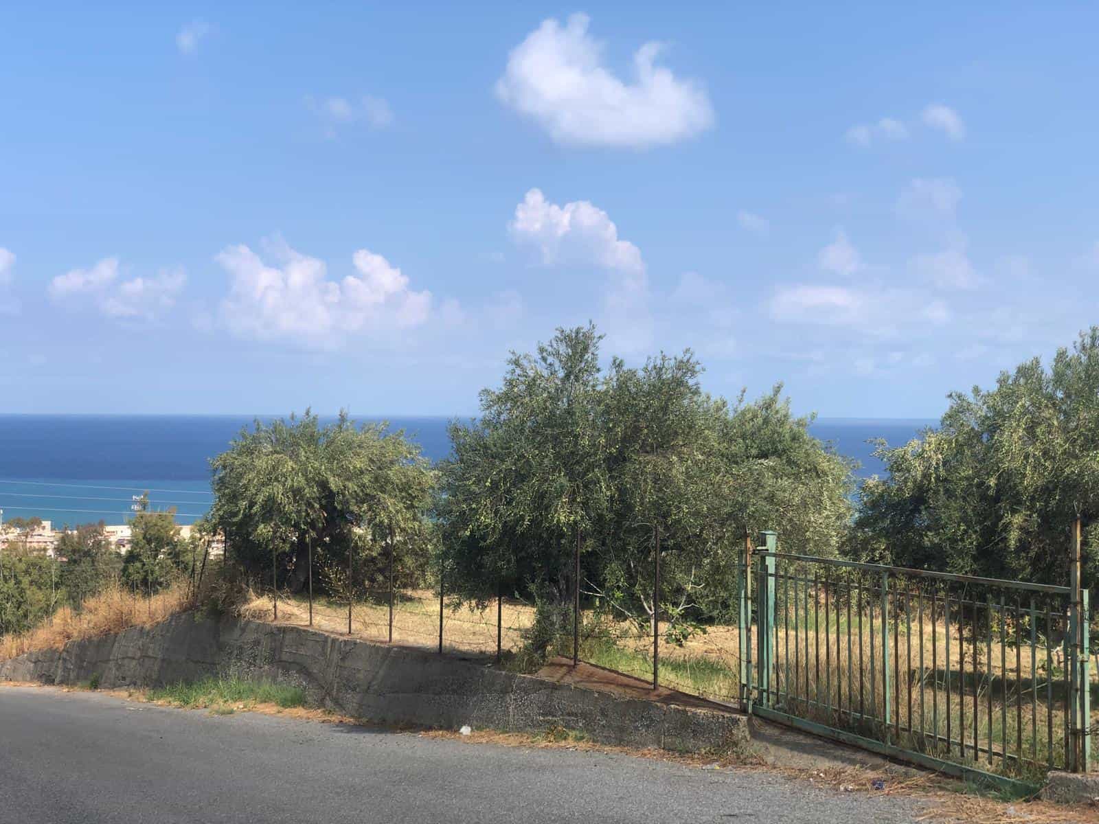 Vasta vista mare con cielo blu e nuvole bianche, sullo sfondo il mare e colline verdi, tone di natura e tranquillità in un contesto di terreno edificabile con alberi e recinzione in ferro.
