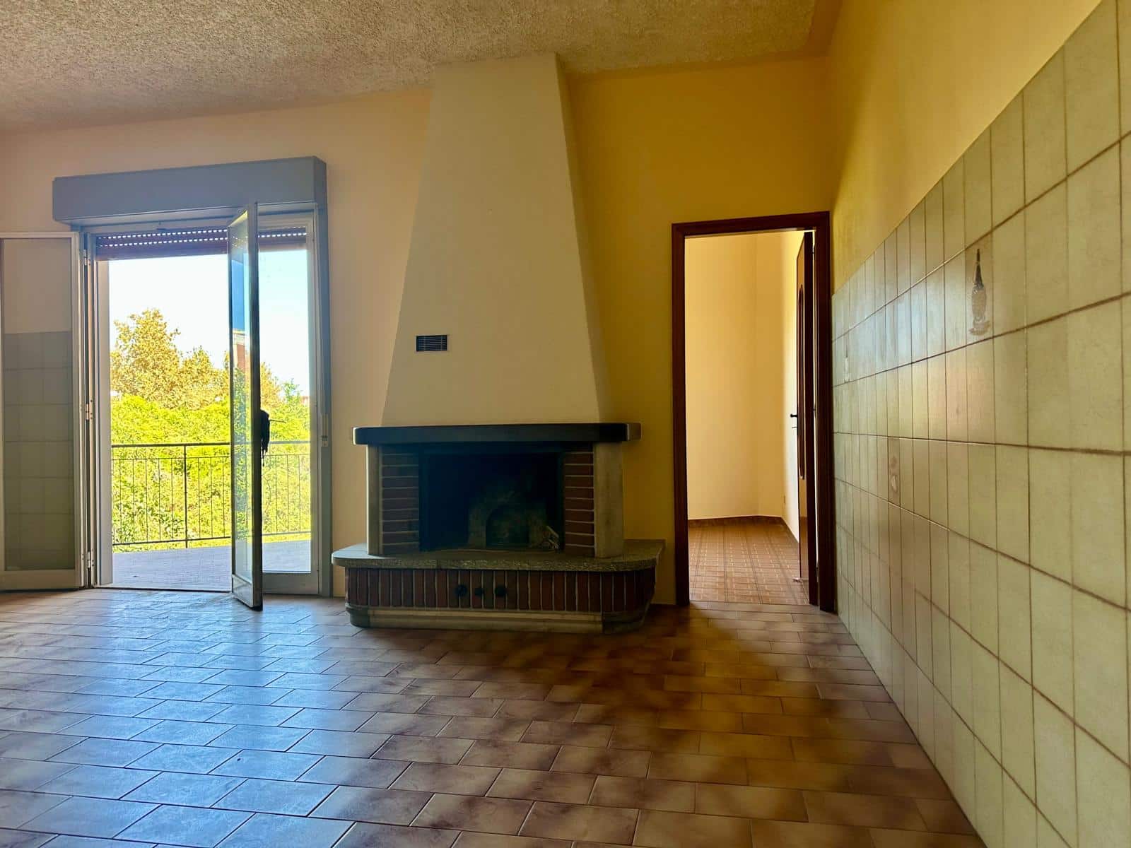 Balcone con porta finestra e camino in soggiorno, vista verde, ambiente spazioso e luminoso, ideale per case e immobili in vendita o affitto.