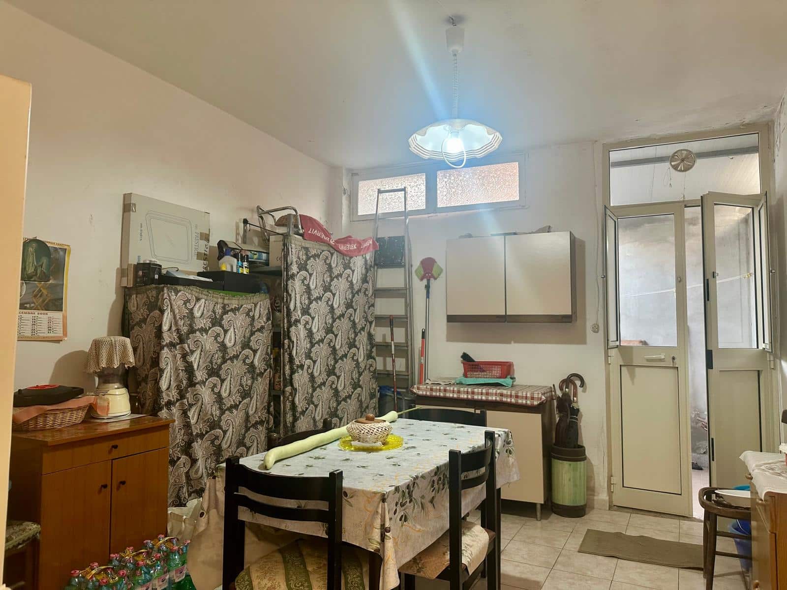 Stanza abitabile con tavolo, sedie, armadio, scaffali, e accessori vari in stile semplice, illuminata da luce naturale, con balcone e ingresso esterno visibili, situata in un appartamento residenziale.