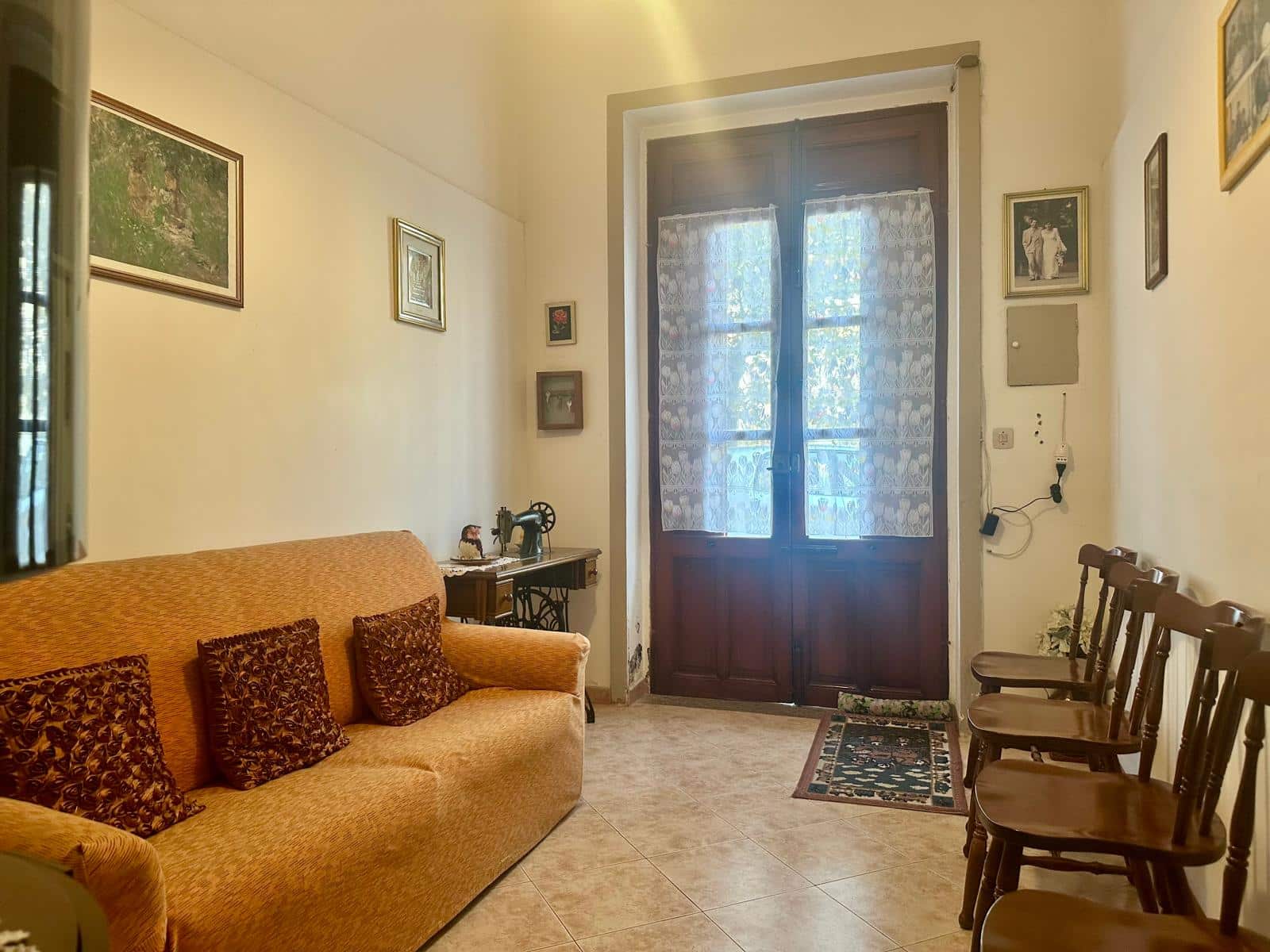 Antico soggiorno con divano in velluto arancione, sedie di legno lungo parete, finestre con tende leggere, decorazioni vintage e macchina da cucire, ambiente accogliente e tradizionale.