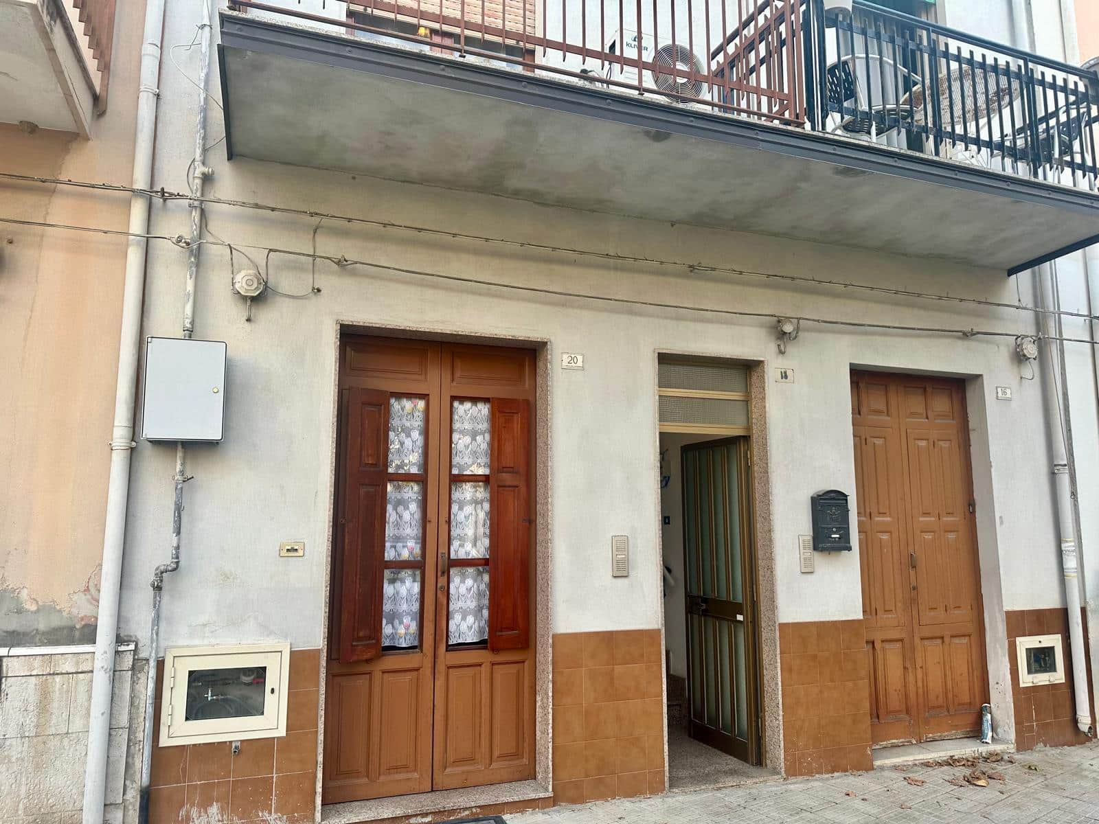 Altezza casa con ingresso, portoncino di legno con vetri decorativi, porta secondaria, facciata in muratura chiara, balcone al primo piano con ringhiera in ferro e oggetti da balcone, dettagli di impiantistica esterna e grate di sicurezza.
