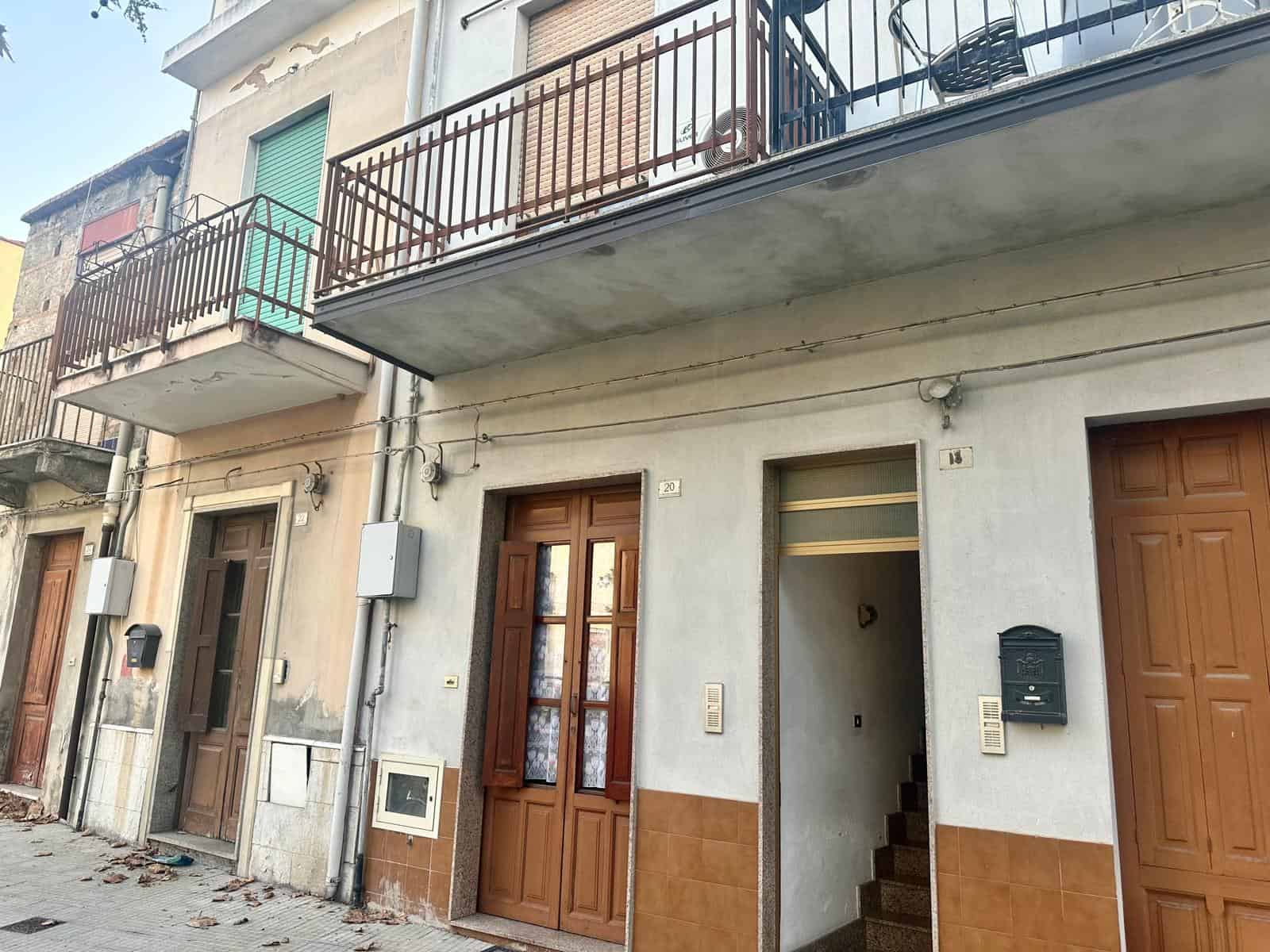 Scala di accesso e ingresso di un edificio residenziale con balcone e serramenti in legno, situato in zona urbana con pavimentazione in pietra e dettagli di manutenzione esterna.