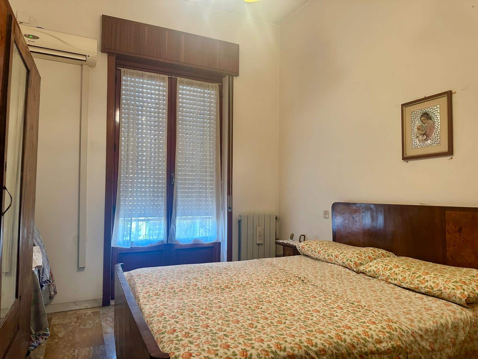 Letto matrimoniale in camera da letto con mobili in legno e tende alle finestre, ambiente luminoso e accogliente con aria condizionata, ideale per soluzioni di affitto o vendita immobiliare.