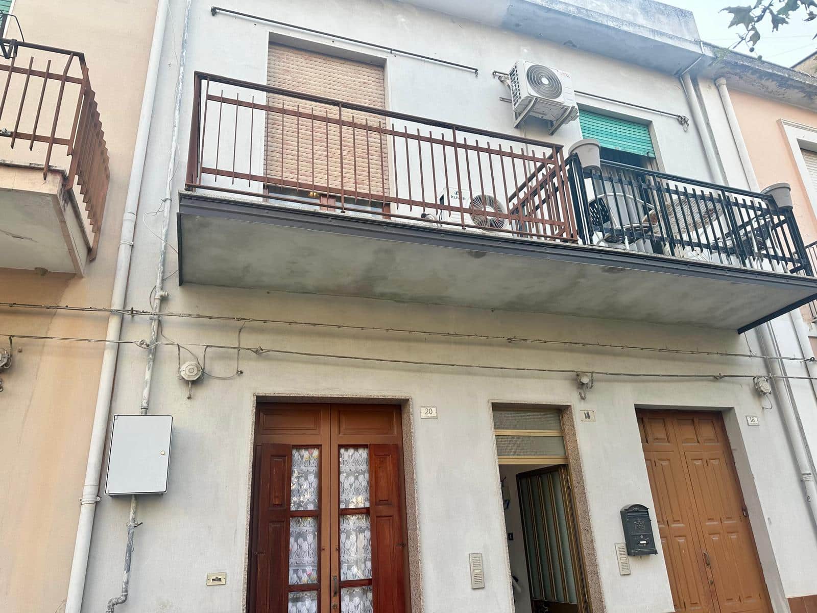 Balcone di un appartamento residenziale con ringhiera in metallo, aria condizionata e serrande abbassate, vista strada urbana, edificio in mattoni e cemento, stile abitativo italiano.
