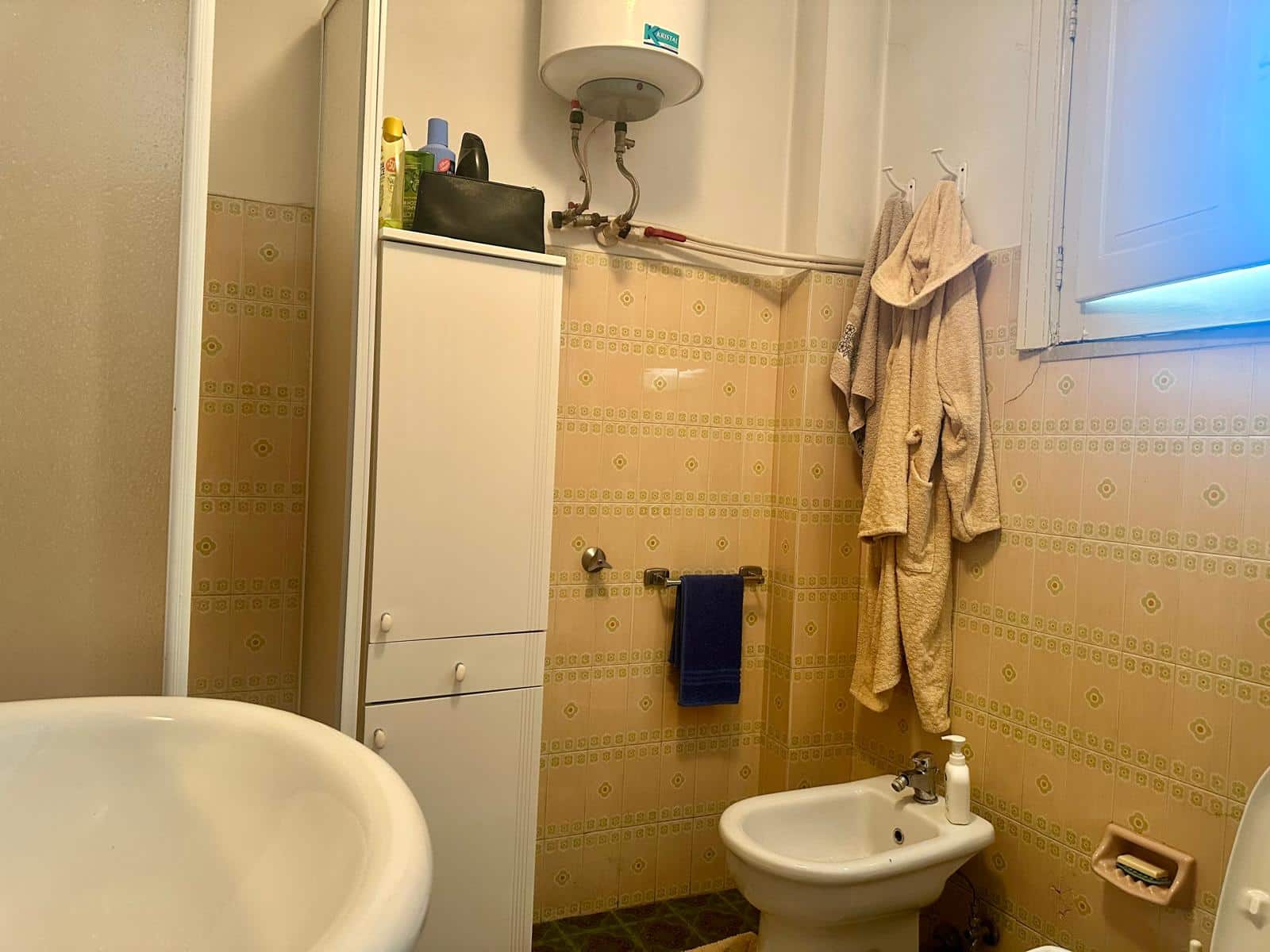 Armadio bianco in bagno con accessori e asciugamani, pareti rivestite in piastrelle gialle e presenza di bidet e spazio per prodotti di pulizia, ambiente funzionale e compatto.