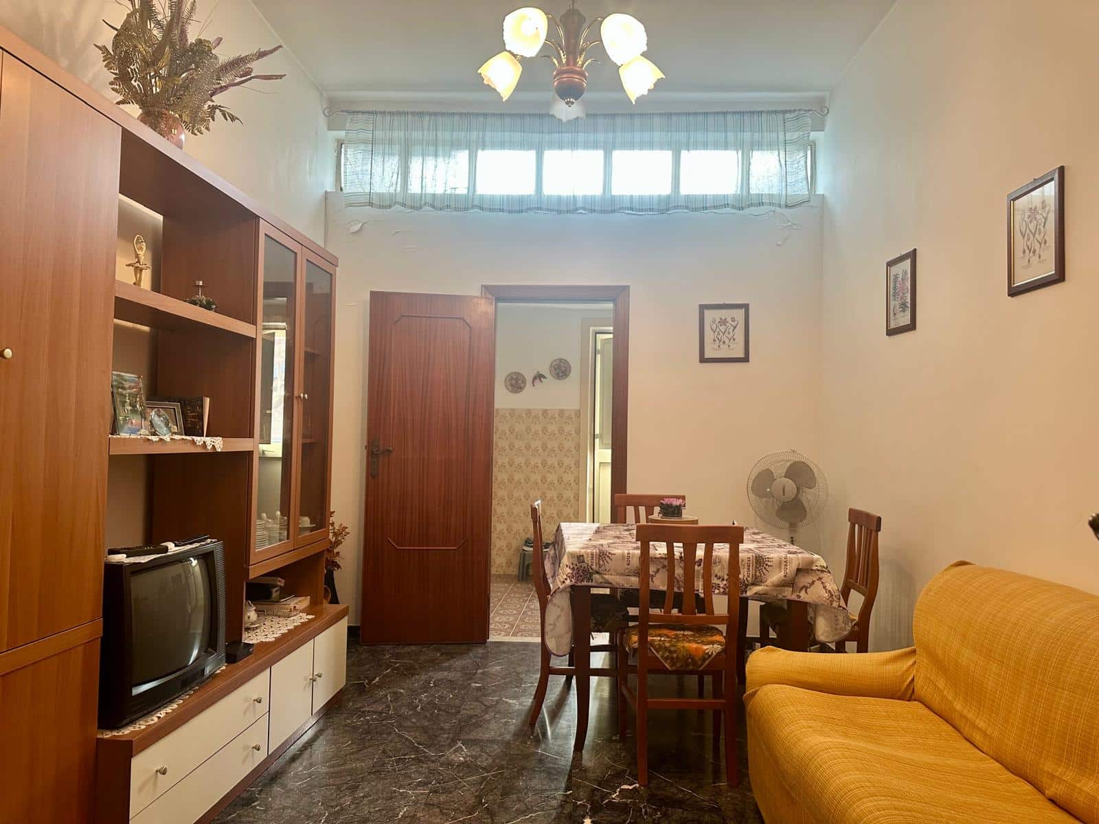 Divano giallo, soggiorno con tavolo da pranzo e mobile porta TV in stile vintage, pareti decorate con quadri, cortile interno, illuminazione a soffitto, atmosfera accogliente e tradizionale.