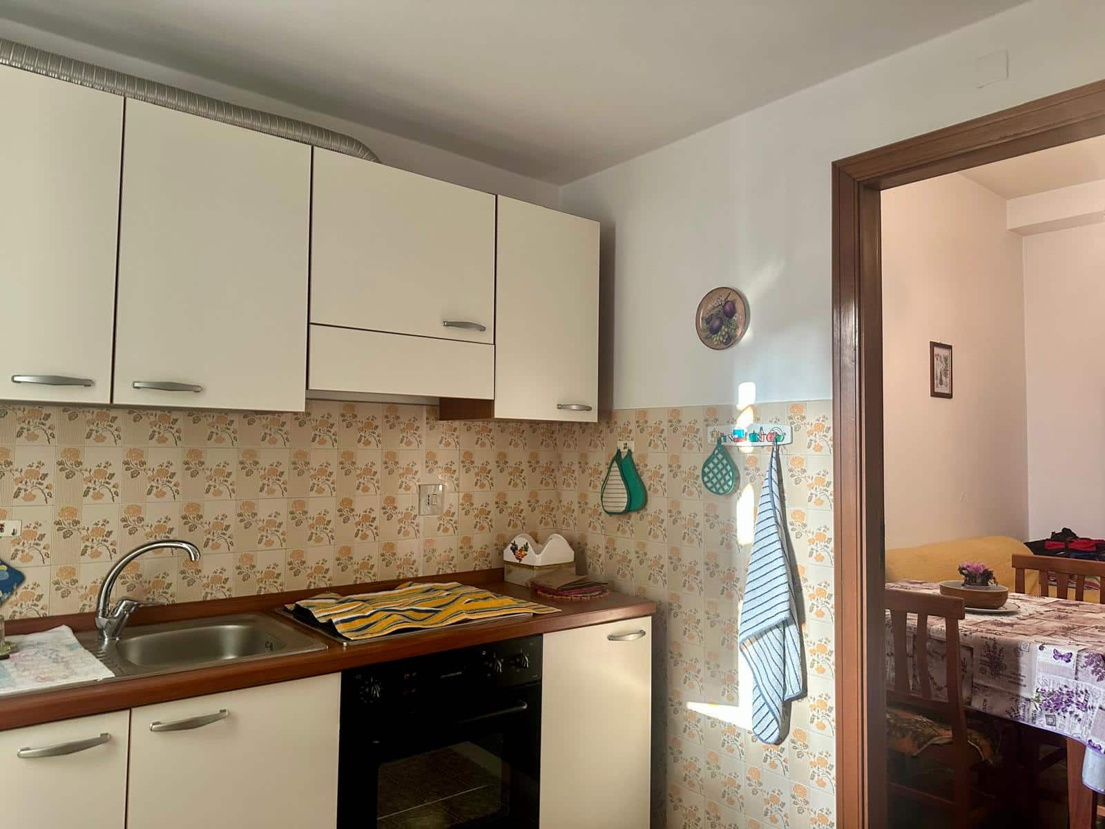Fondo cucina semplice e funzionale con ante bianche e piastrelle floreali, vista su sala da pranzo con tavolo e sedie, in un appartamento accogliente NextCasa.
