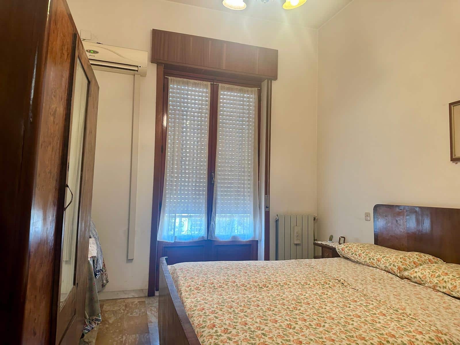 Camera da letto con letto matrimoniale, armadio in legno, finestra con tende a rullo, aria condizionata, arredamento classico, pareti bianche, bagno vicino, atmosfera accogliente e luminosa, ideale per case in vendita o affitto.