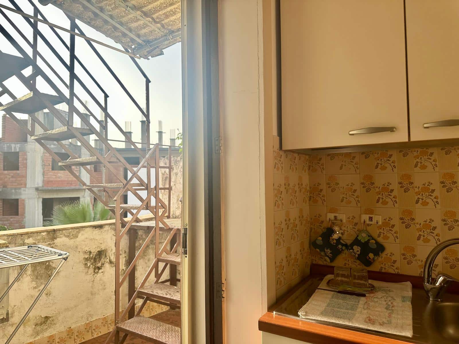 Balcone con scala in ferro battuto e vista sul cielo e sulle facciate delle case vicine, accessibile dalla cucina con dettagli di piastrelle vintage e lavabo.
