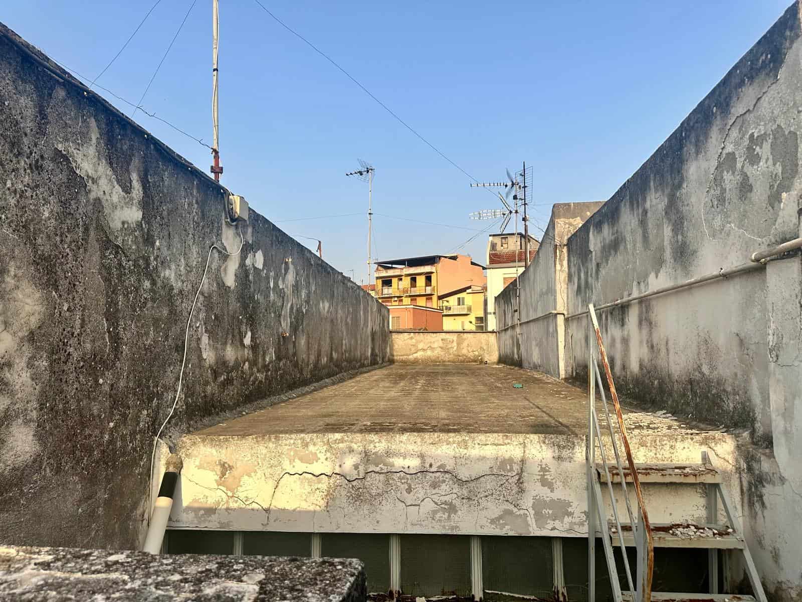 Tetto panoramico con veduta su edifici urbani, superficie in cemento, pareti nere e grigie, staccionata in metallo arrugginita, scale di accesso in ferro arrugginito, ambiente esterno di un condominio, ascensore per tetto, altro edificio visibile sullo sfondo.