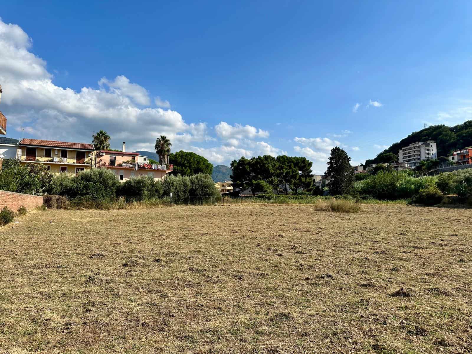 Campo edificabile con case e villette in background e cielo azzurro con nuvole, opportunità di investimento immobiliare in zona residenziale vicino alla natura.