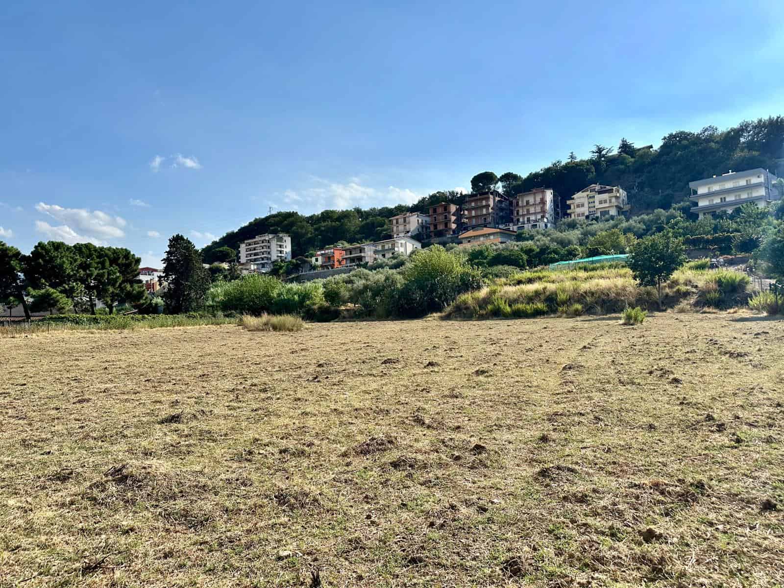 Prato di terra con case moderne sulla collina, vegetazione rigogliosa, cielo blu soleggiato, zona residenziale italia.