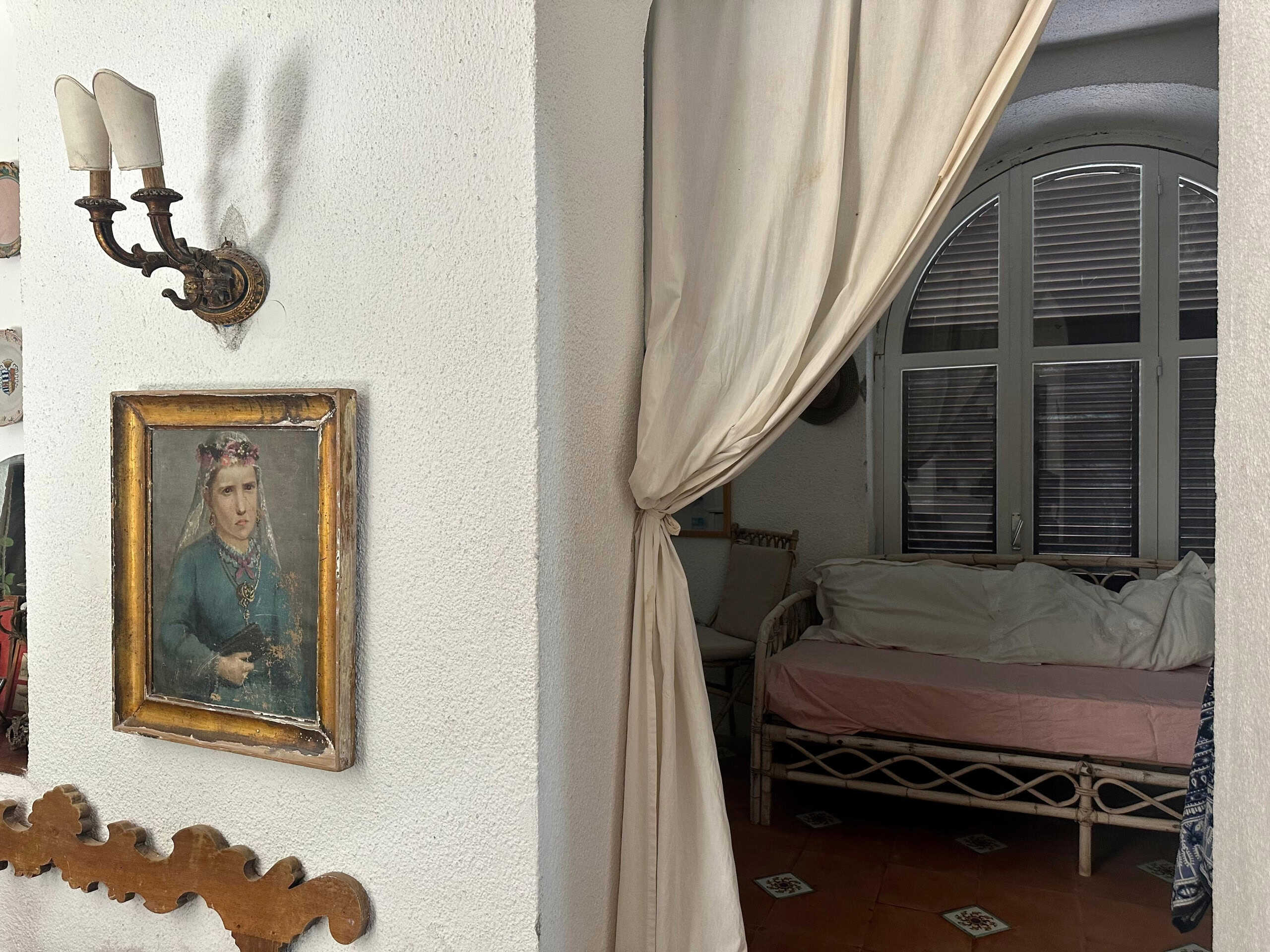 Vecchio letto in vimini con lenzuola bianche, finestra ad arco con persiane di legno, tenda beige che divide le stanze, arredamento rustico e decorazioni antiche, stanza da letto in stile classico, ambiente accogliente e luminoso, immagini di interni per case rustiche e di charme, arredamento vintage, NextCasa.