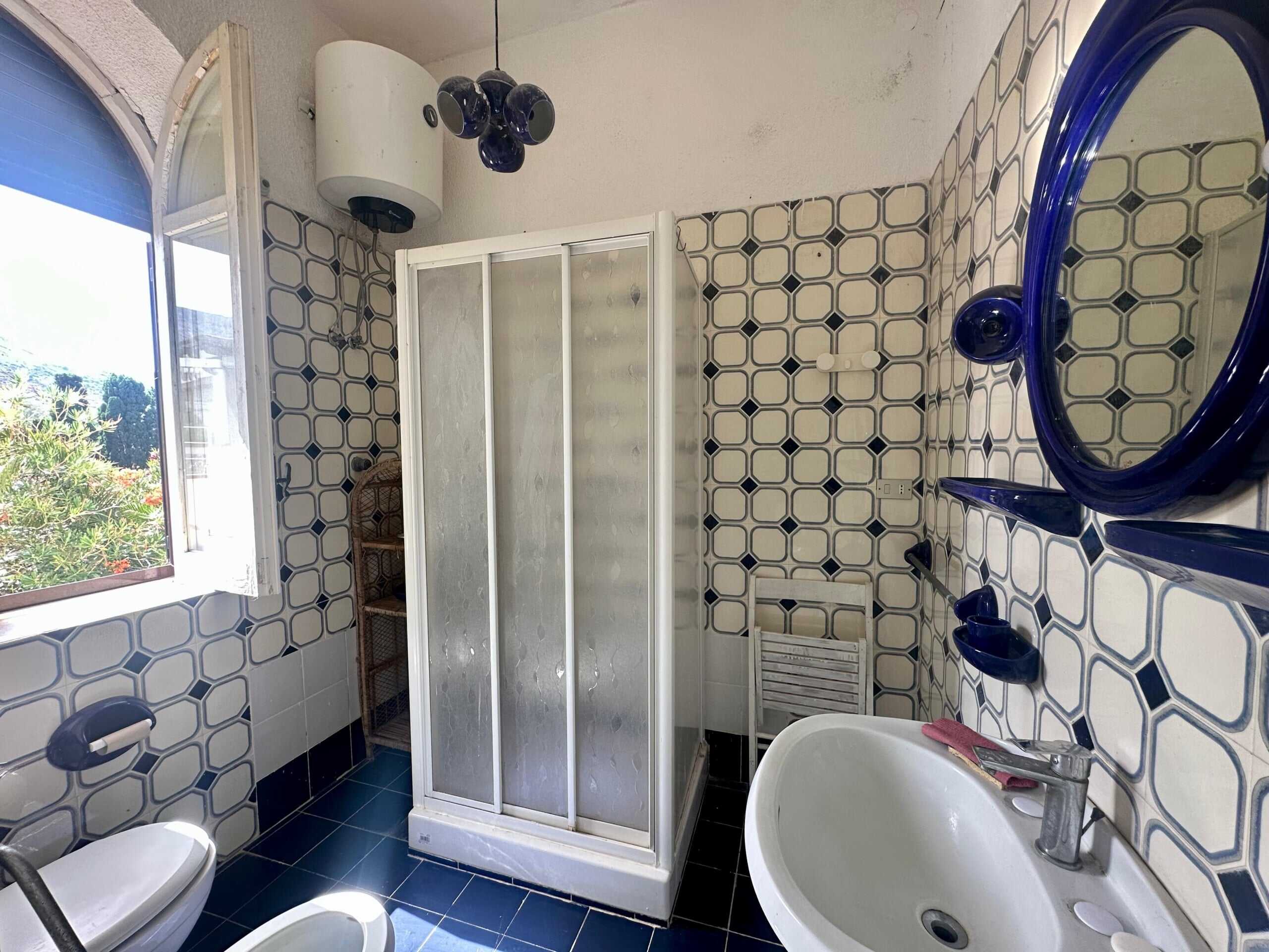 Doccia in bagni ristrutturati con piastrelle decorative, finestra con vista sul verde, lavabo elegante e specchio blu, mobili funzionali, ideale per case di charme e comfort moderno.