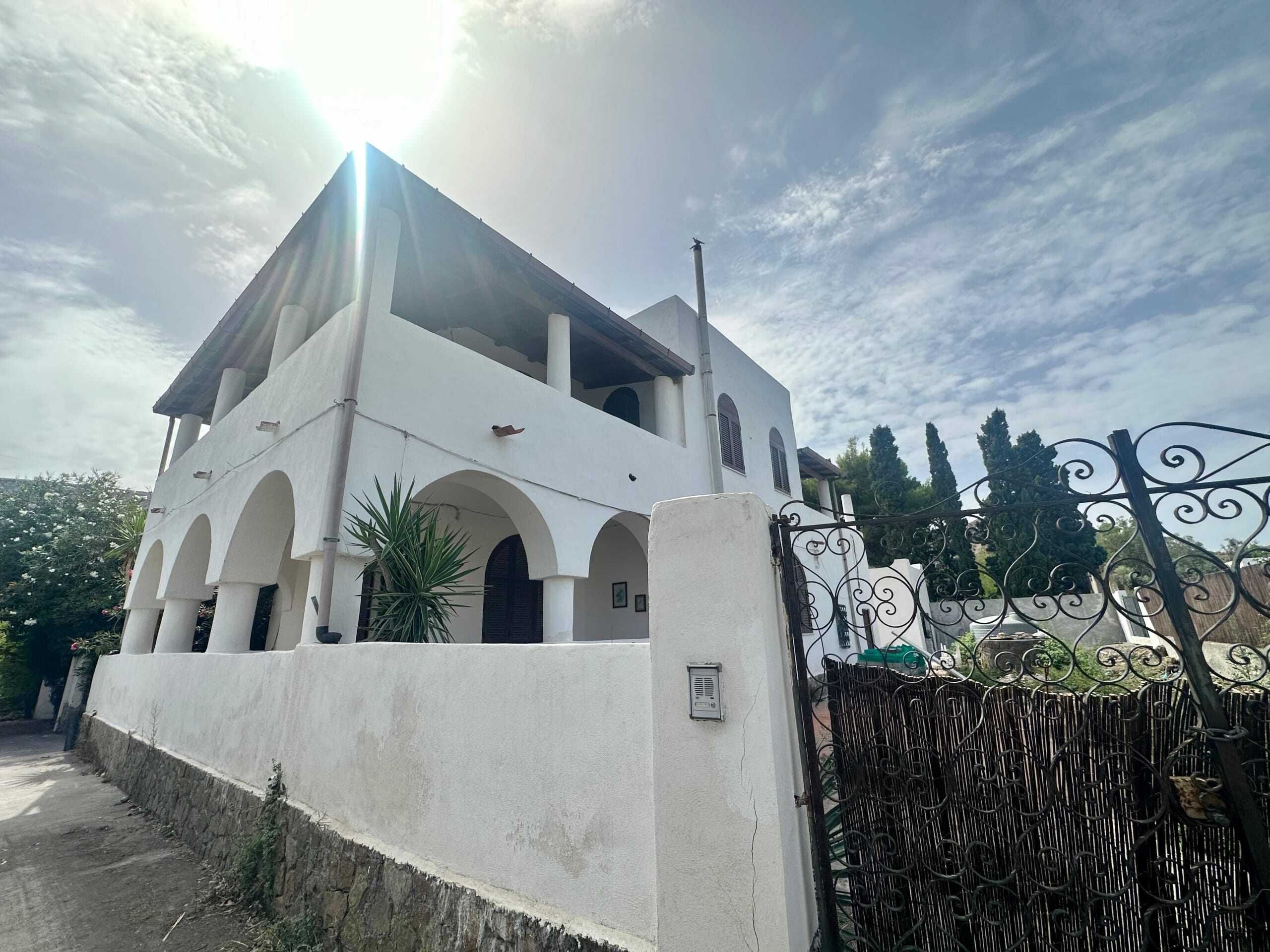 Villa bifamiliare con pareti bianche, portico e giardino, stile mediterraneo, in Sicilia, con ingresso in ferro battuto, zona verde e atmosfera accogliente, ideale per residenza estiva o investimento immobiliare.