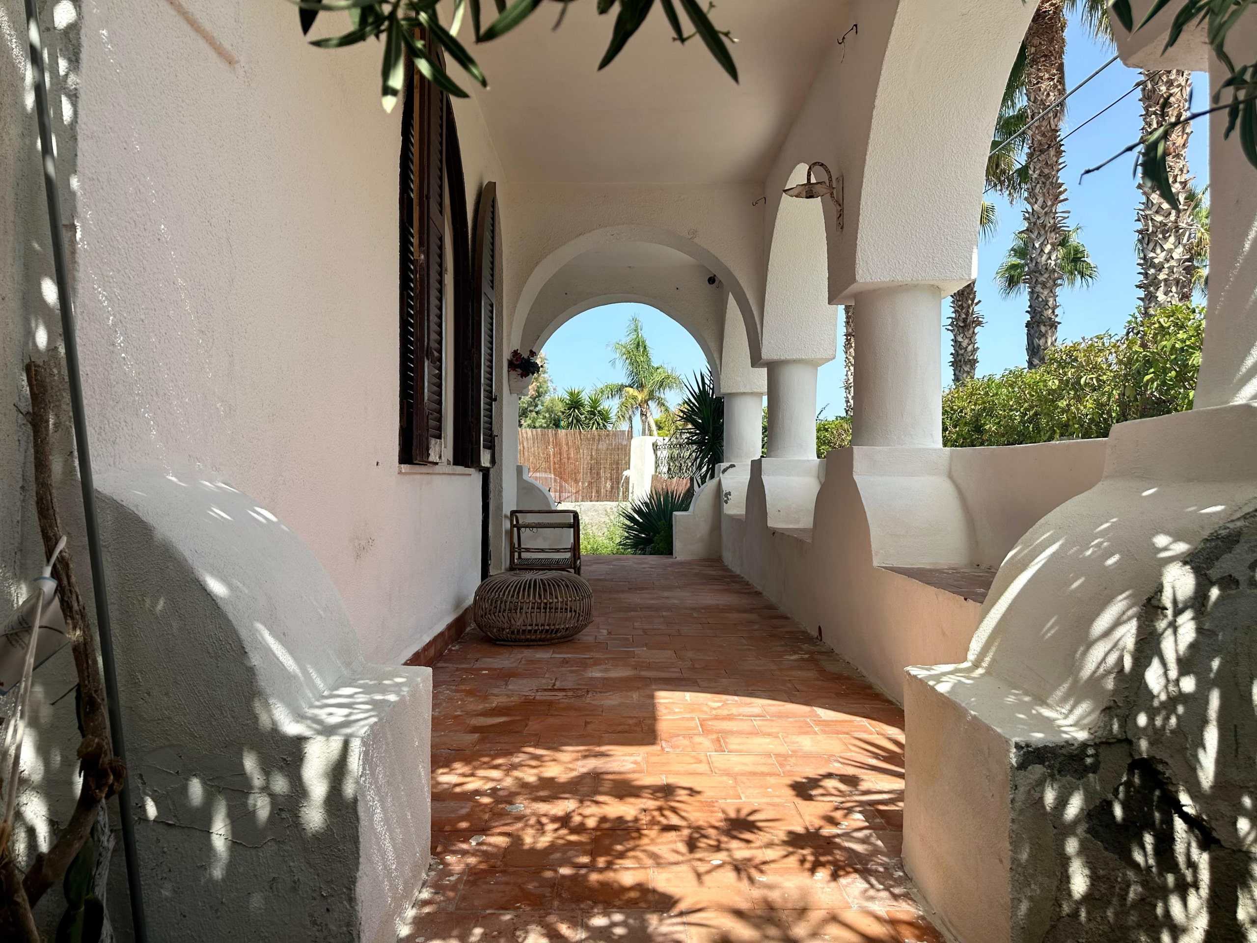 Spazio esterno con terrazza in stile mediterraneo, pareti bianche, archi e colonne, panchine in pietra e piante tropicali, ideale per relax e outdoor living.