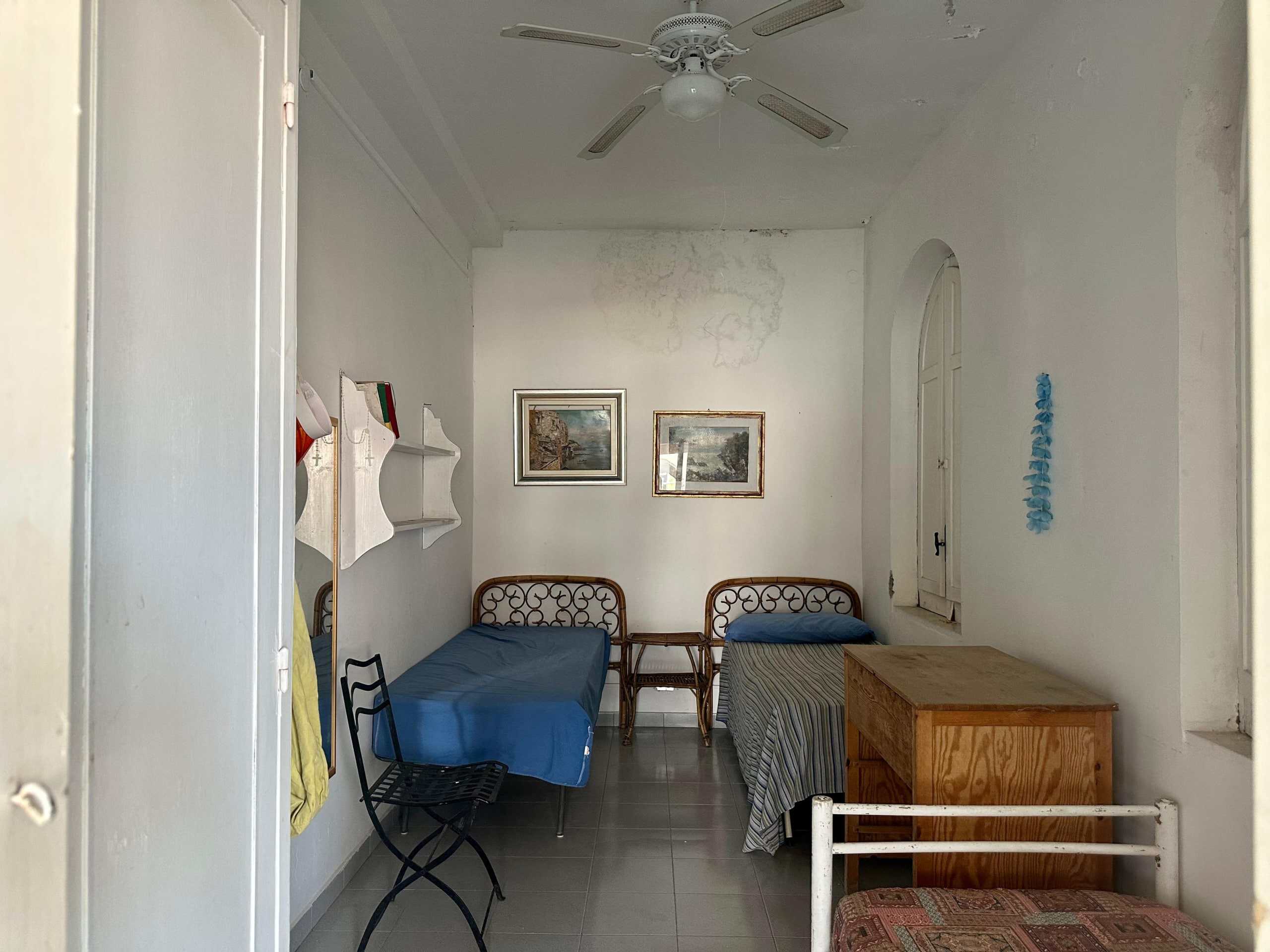 Vecchio camera da letto con mobili vintage, pareti bianche, due letti singoli con testiere in ferro battuto, quadri alle pareti e finestra ad arco, atmosfera rustica e semplice.