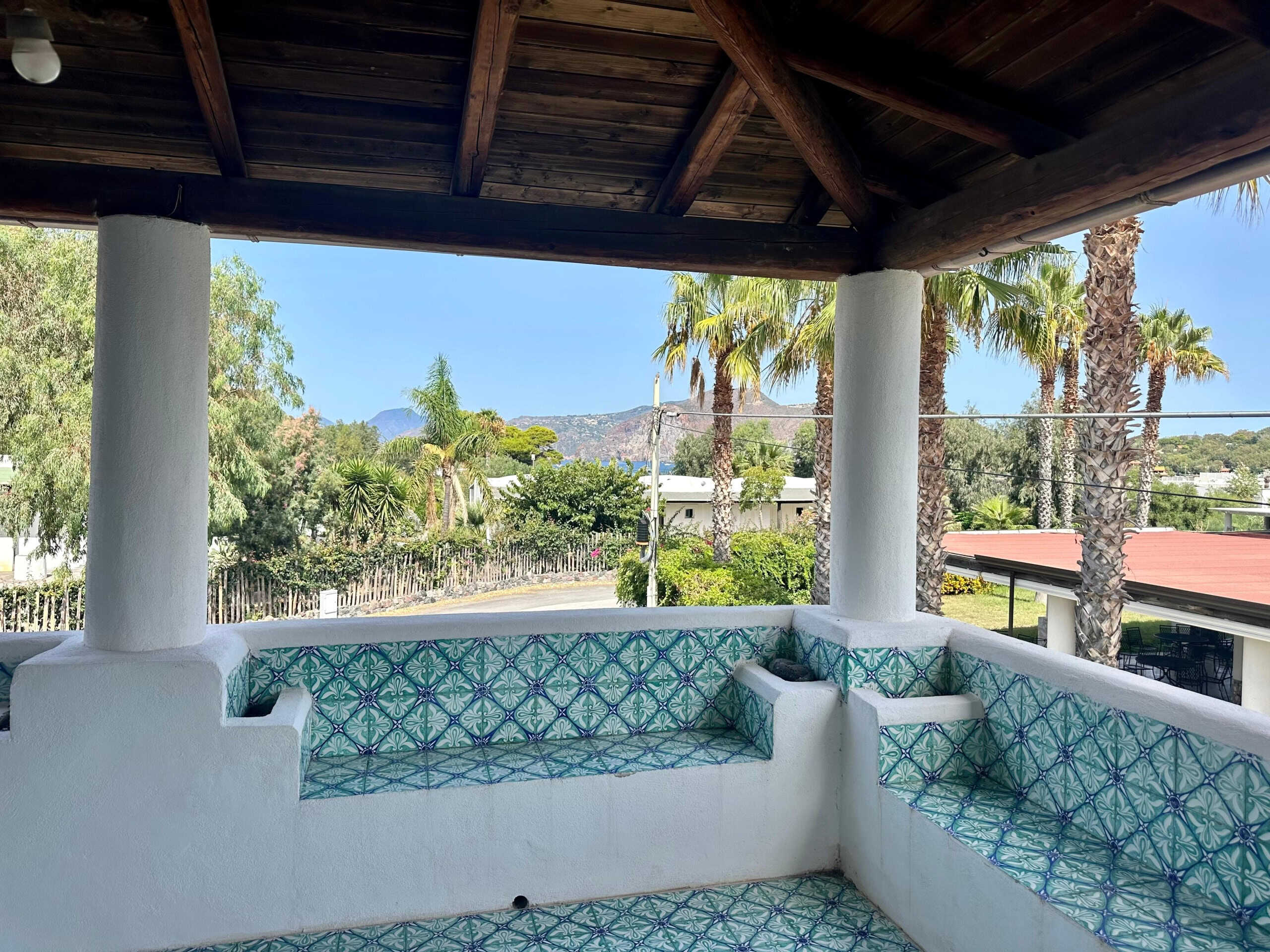 Vista sulla terrazza con panche decorate in stile mediterraneo, palmizi e vegetazione tropicale, con montagna sullo sfondo, in una zona residenziale soleggiata.