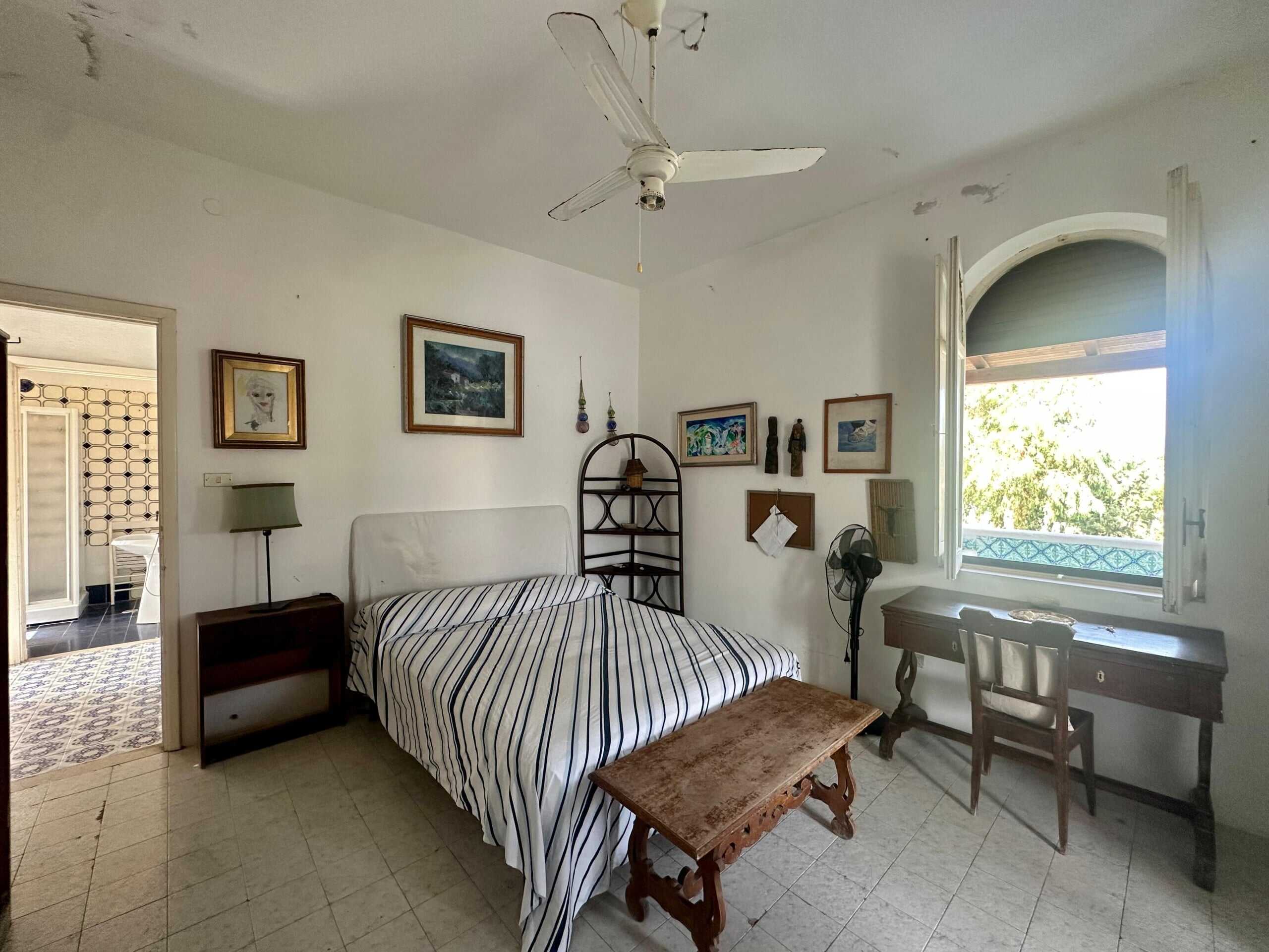 Letto matrimoniale con biancheria a righe, stanza da letto con pareti bianche, quadro alle pareti e angolo studio con scrivania e sedia, finestra aperta con vista su albero, ventilatore e lampada da tavolo.