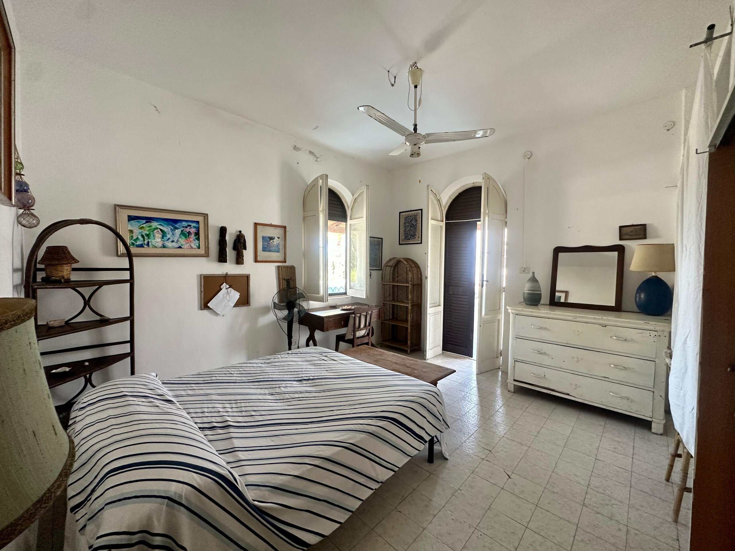 Vecchio appartamento con mobili rustici e decorazioni artistiche, luminosa stanza da letto con doppio letto, finestre con persiane, armadi in legno e pareti bianche, ambiente accogliente e spazioso.