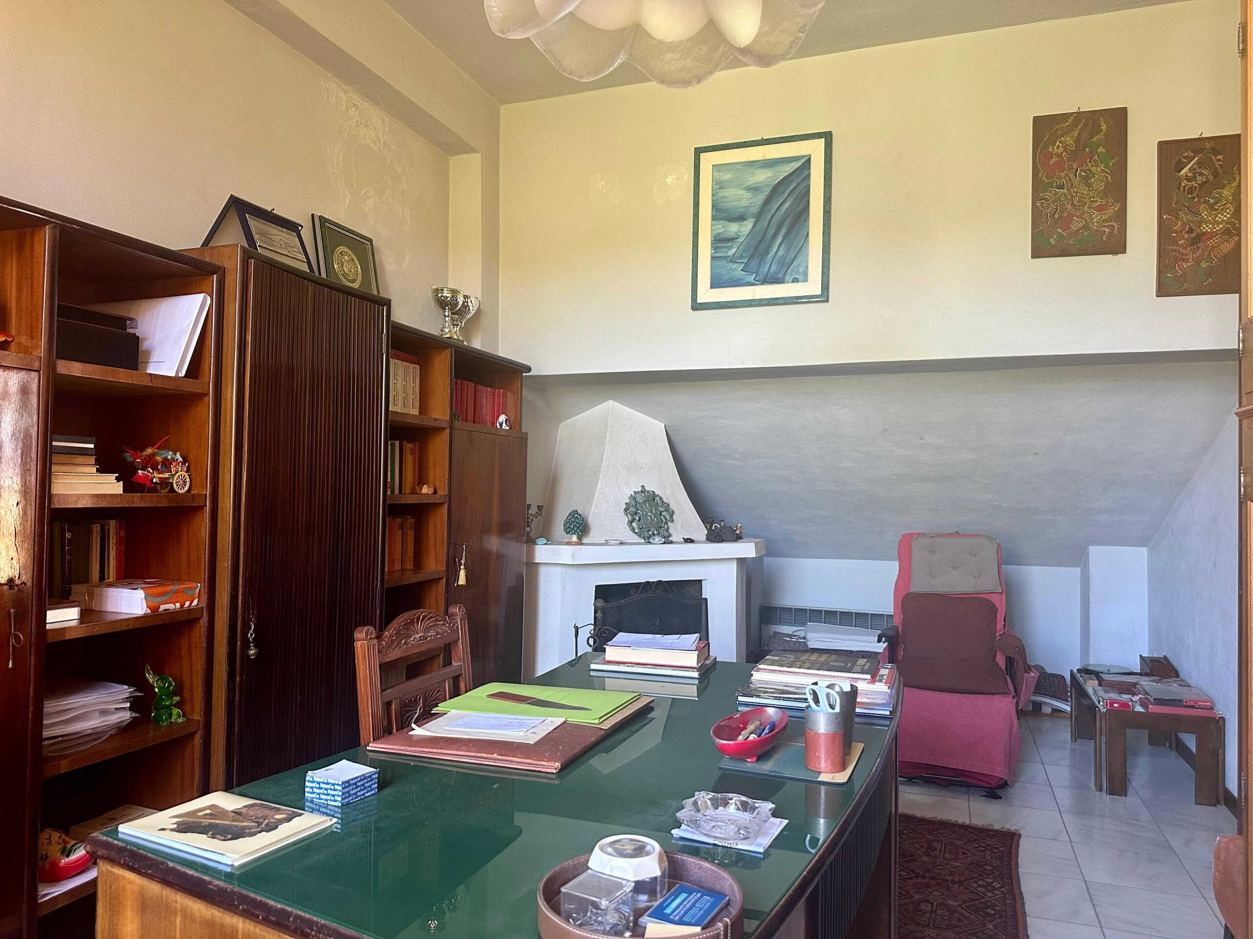 Archivio immobili, ufficio domestico con mobili in legno, libri, scrivania con documenti e oggetti decorativi, camino, sedie e tende, ambiente accogliente e funzionale.