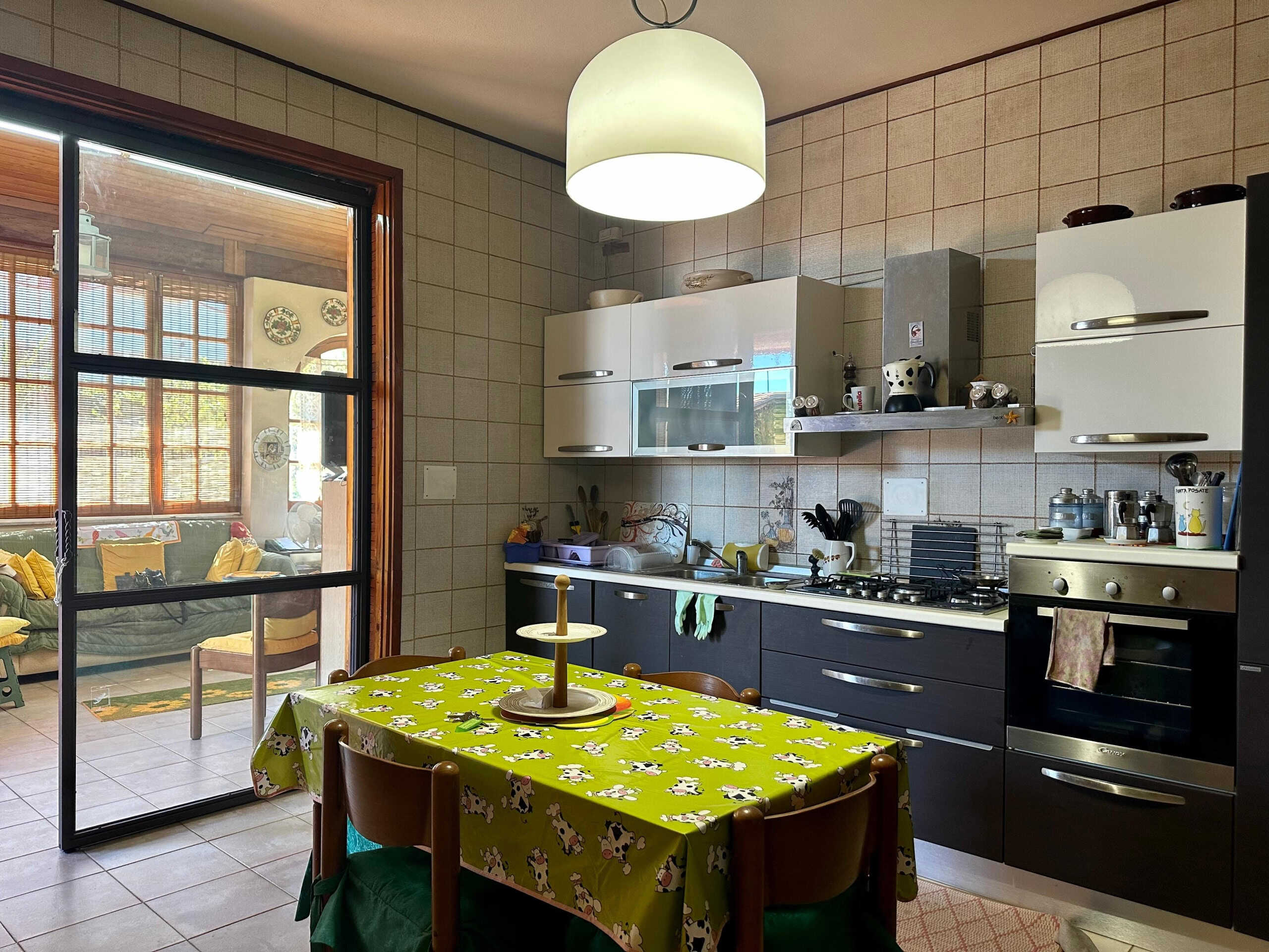 Atmosfera accogliente e funzionale in cucina: arredi moderni, tavolo con tovaglia vivace, accessori pratici e apertura sul salotto con verità a vetri, ideale per vivere e condividere momenti familiari.