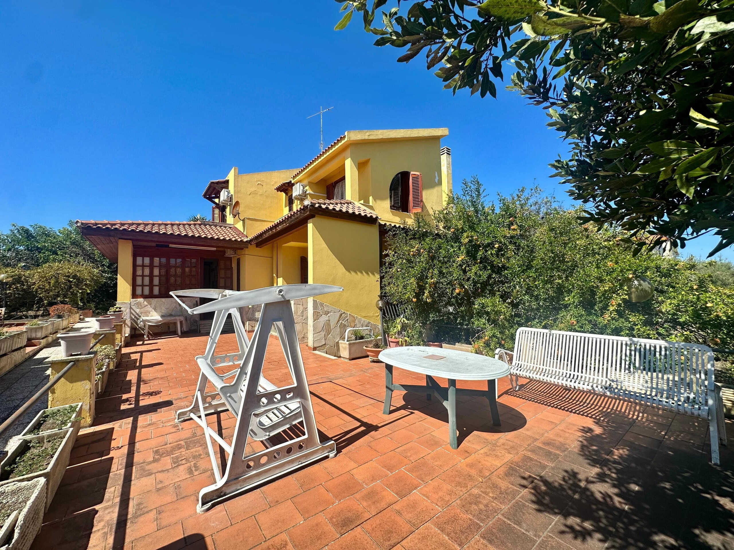 Spazio esterno di casa con terrazza soleggiata, mobili da giardino, piante e vista su un edificio colorato, ideale per relax e convivialità.