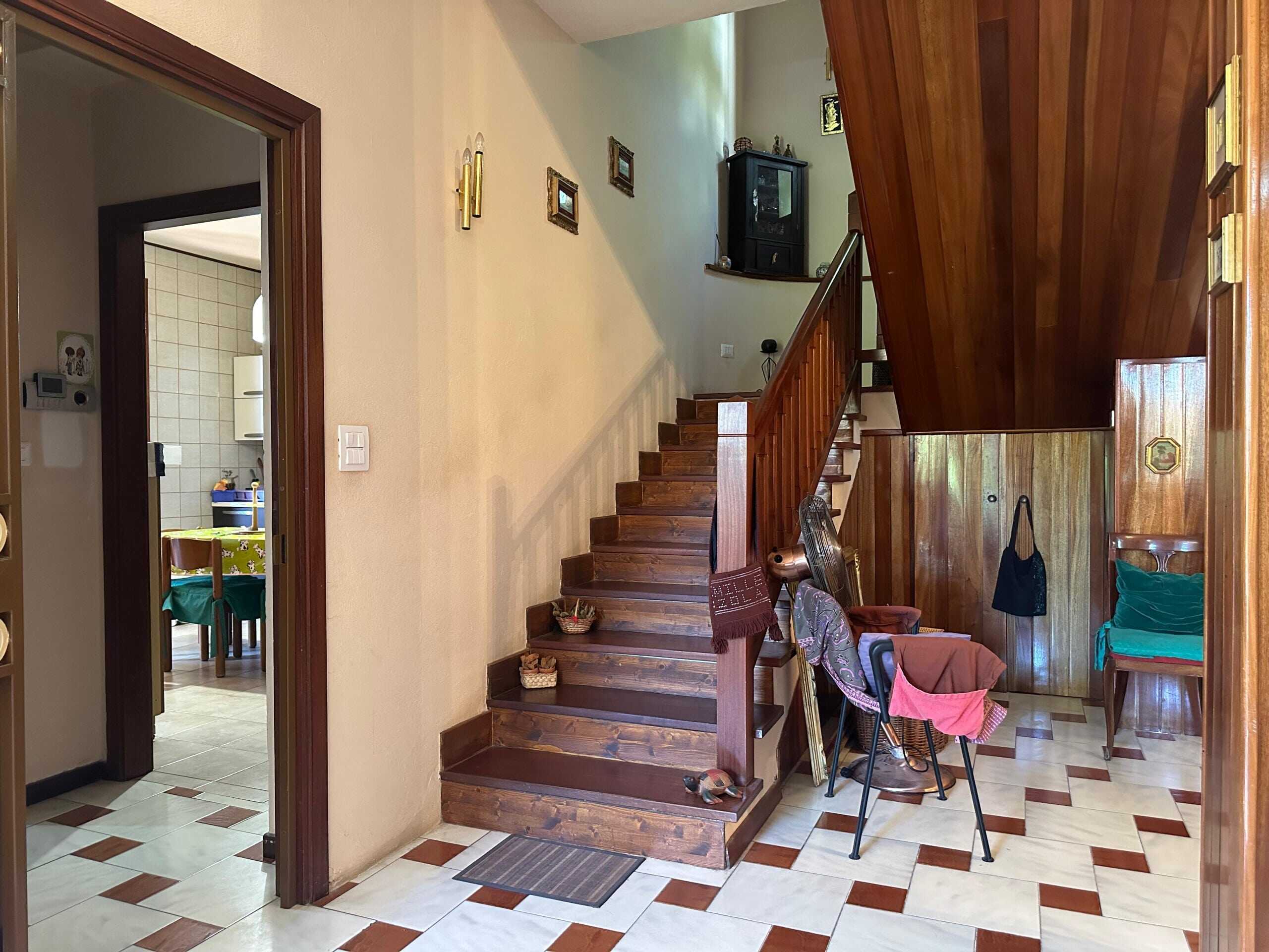 Scalinata in legno, ingresso di casa con pareti beige e pavimento in piastrelle bianche e marroni, porta della cucina aperta, oggetti come ceste, ventola e borse, ambiente accogliente e rustico.
