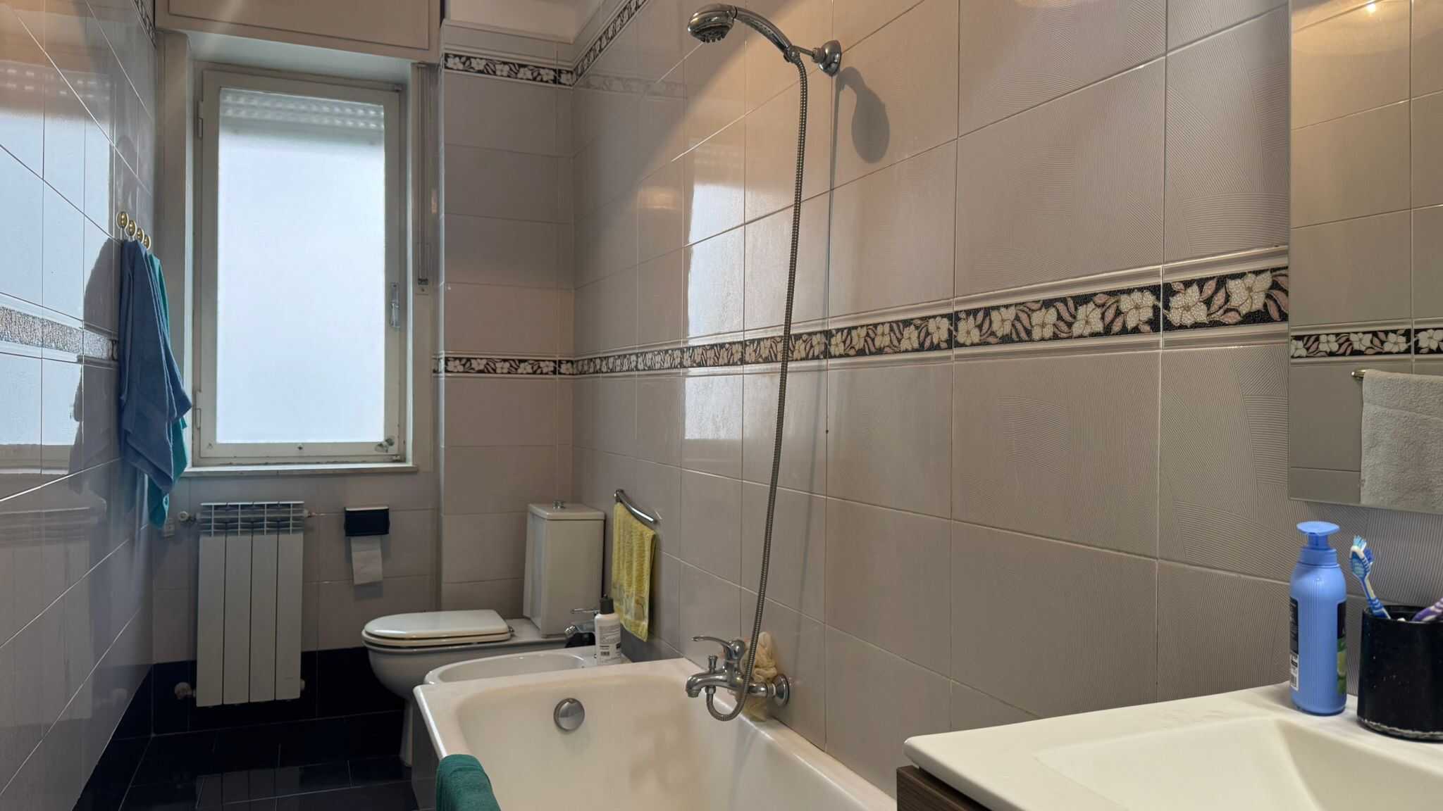 Bagno con vasca e bidet, finestra con tendina, arredo bagno semplice con prodotti per l'igiene, piastrelle beige con decoro floreale, ambiente luminoso e funzionale.