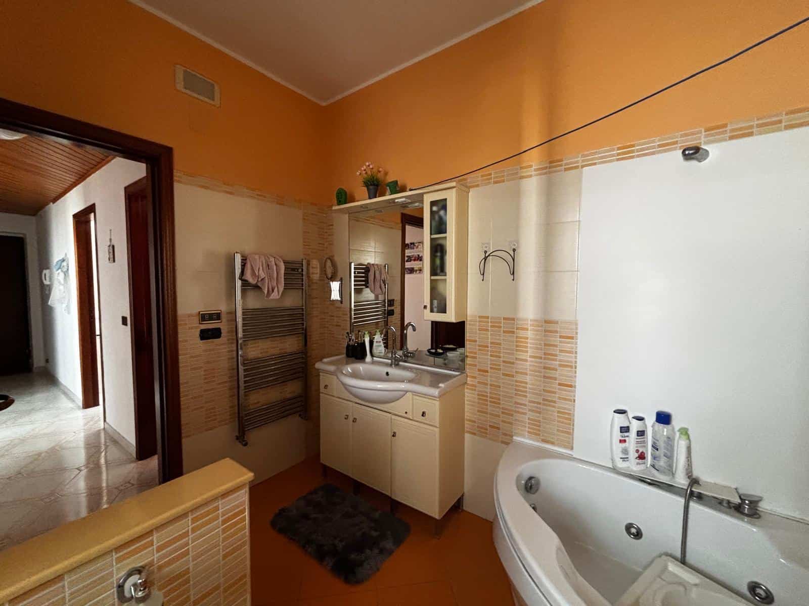 Immagine di un vivace bagno domestico con pareti arancioni e dettagli beige, dotato di vasca angolare, lavabo con mobiletto, asciugamani appesi e mensole con prodotti per l'igiene personale, ideale per abitazioni moderne.
