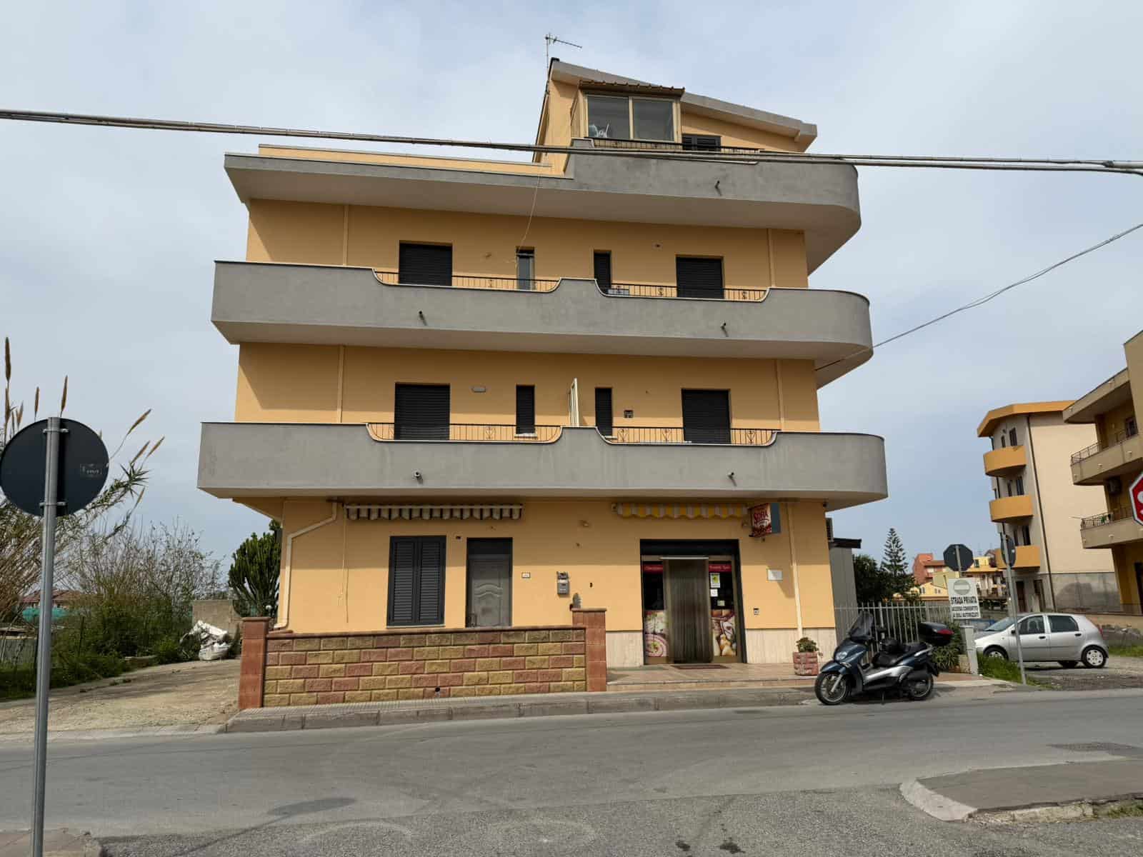 Appartamento moderno con tre piani e balconi ampi, situato in zona urbana con parcheggio e strada trafficata. Edificio residenziale con facciata gialla e dettagli in cemento, vicino a servizi e negozi.