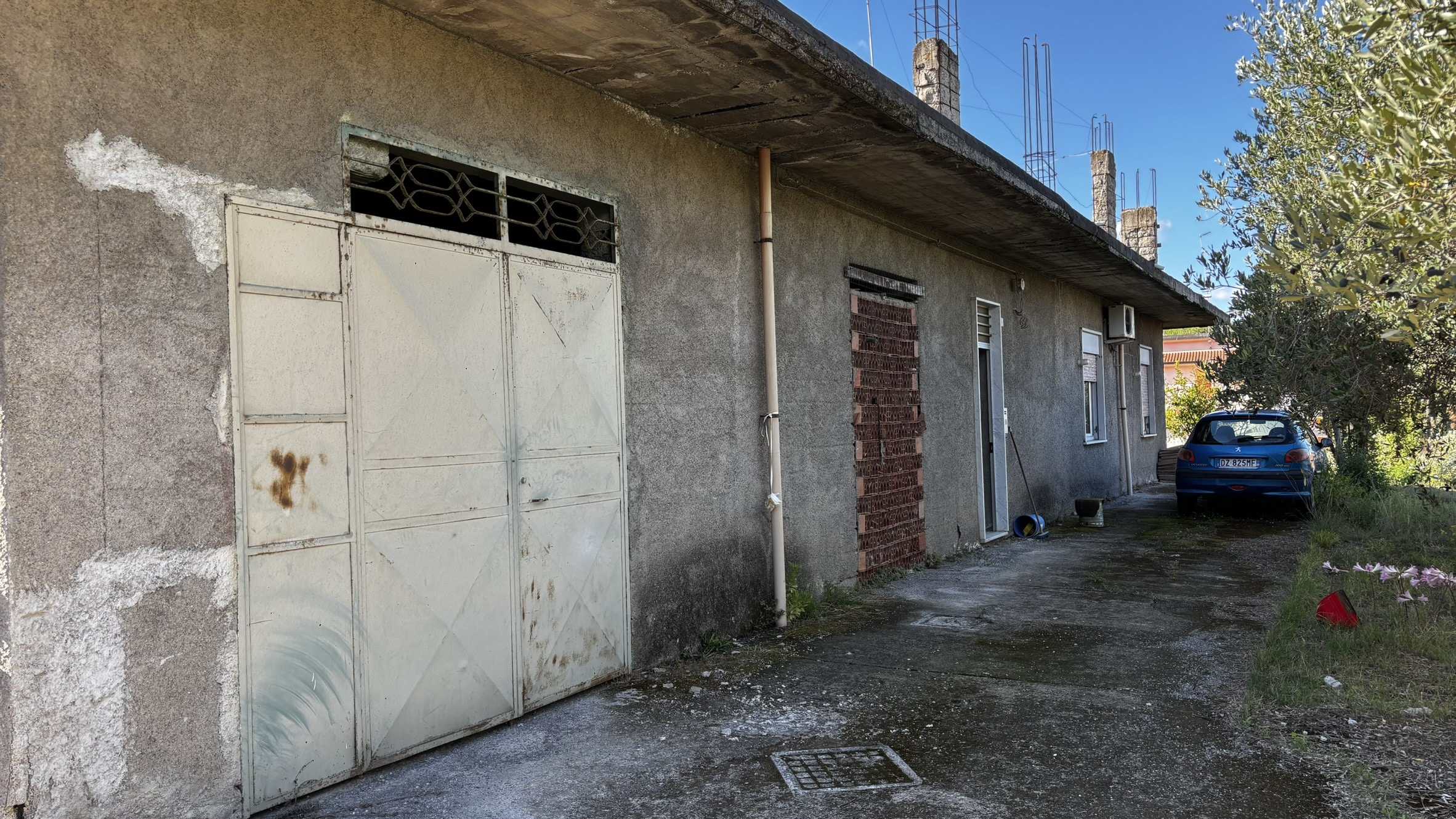 1 Portone garage rustico in mattoni e cemento, esterno di una casa abbandonata, parcheggio con auto, lato cortile in zona residenziale, ambientazione rustica e trascurata, materiale edile visibile, tetto in cemento con pioli di ferro.