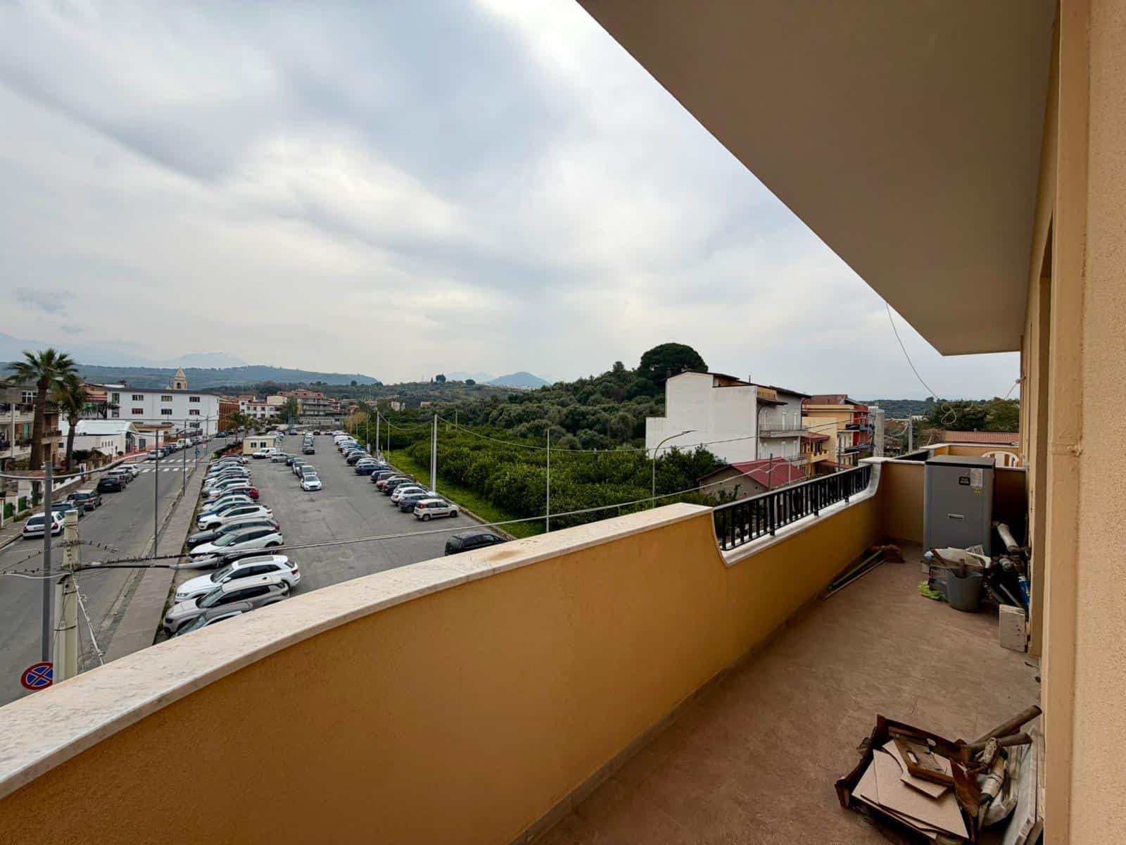 Balcone di un appartamento con vista su strada e aree verdi, con mobili e oggetti di servizio, in un contesto urbano e naturale, in Italia.