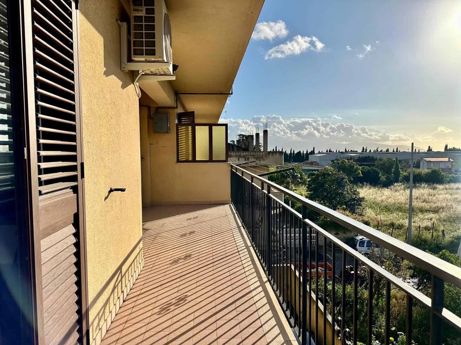 Balcone di un appartamento con vista panoramica sulla città e sulla natura, illuminato dal sole, con ringhiera in metallo e aria condizionata esterna. Ideale per relax e momenti all'aperto.