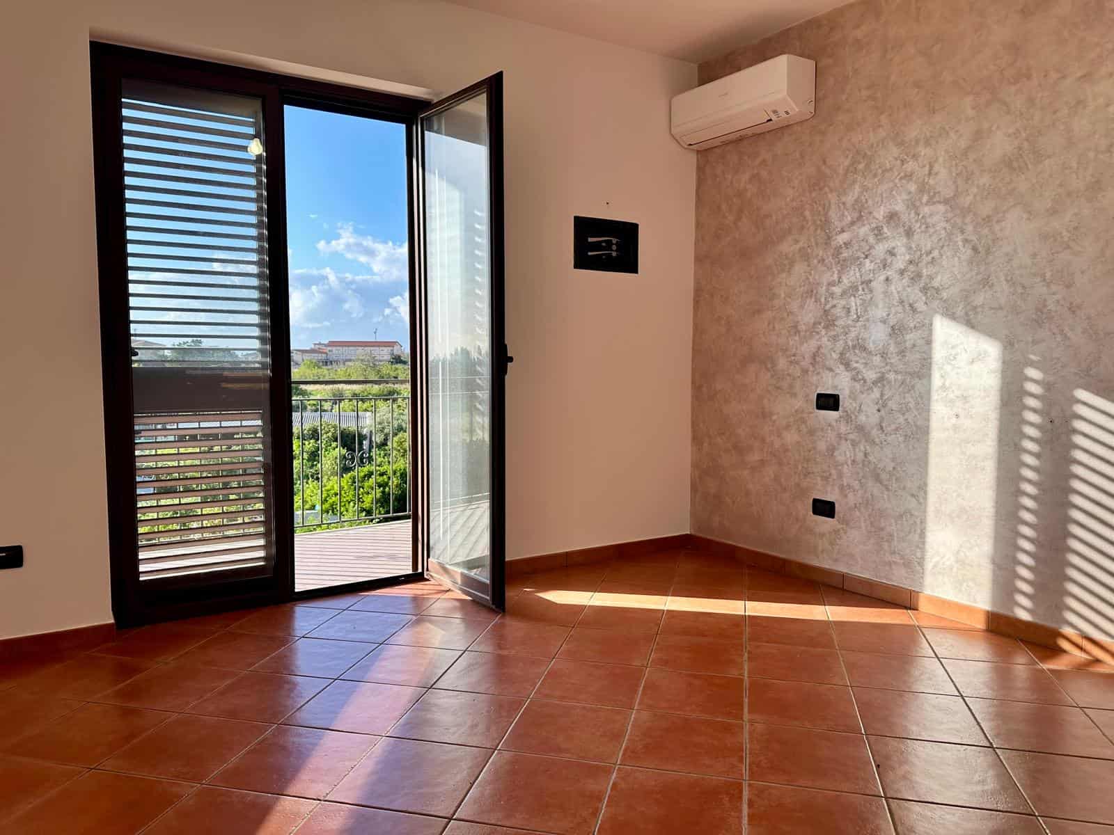 Ampia stanza luminosa con porta finestra che si apre su un balcone, vista sul cielo e sulla natura, aria condizionata moderna, pavimento in ceramica color terracotta, ideale per affitti o acquisti imm.